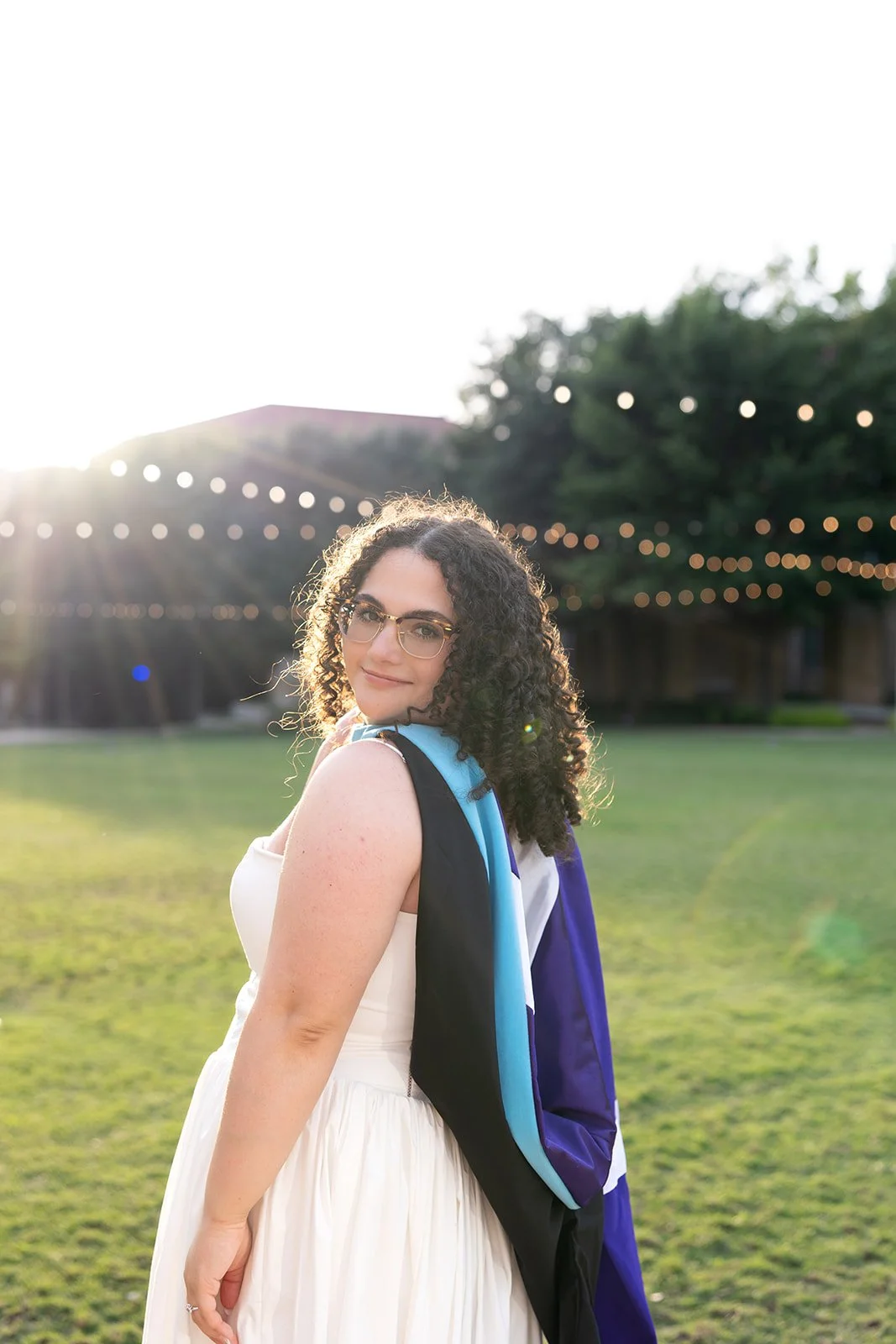 A young woman with curly brown hair, glasses, and a light dress, standing outdoors on a sunny day with a graduation gown over her shoulder. Behind her is a grassy field and string lights with a building and trees in the distance.