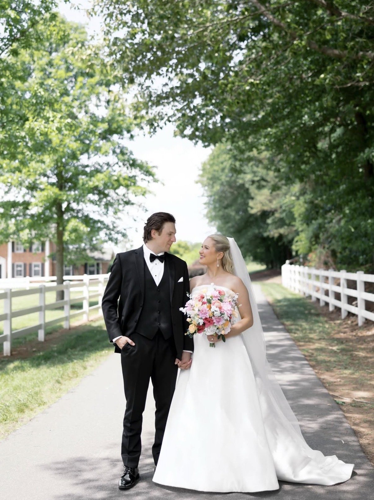 A bride and groom hold hands and look at each other while walking on a path beneath green trees, with white fences on either side, on their wedding day.