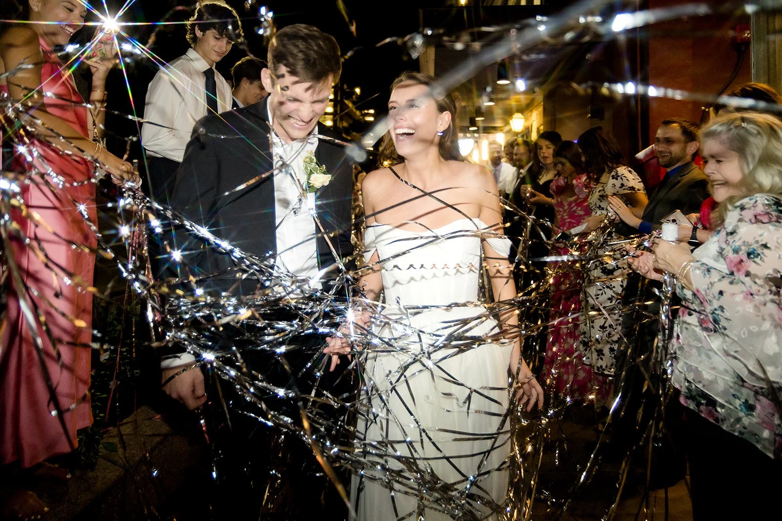 A newlywed couple is celebrating their wedding night, surrounded by friends and family, with confetti and lights in the background.