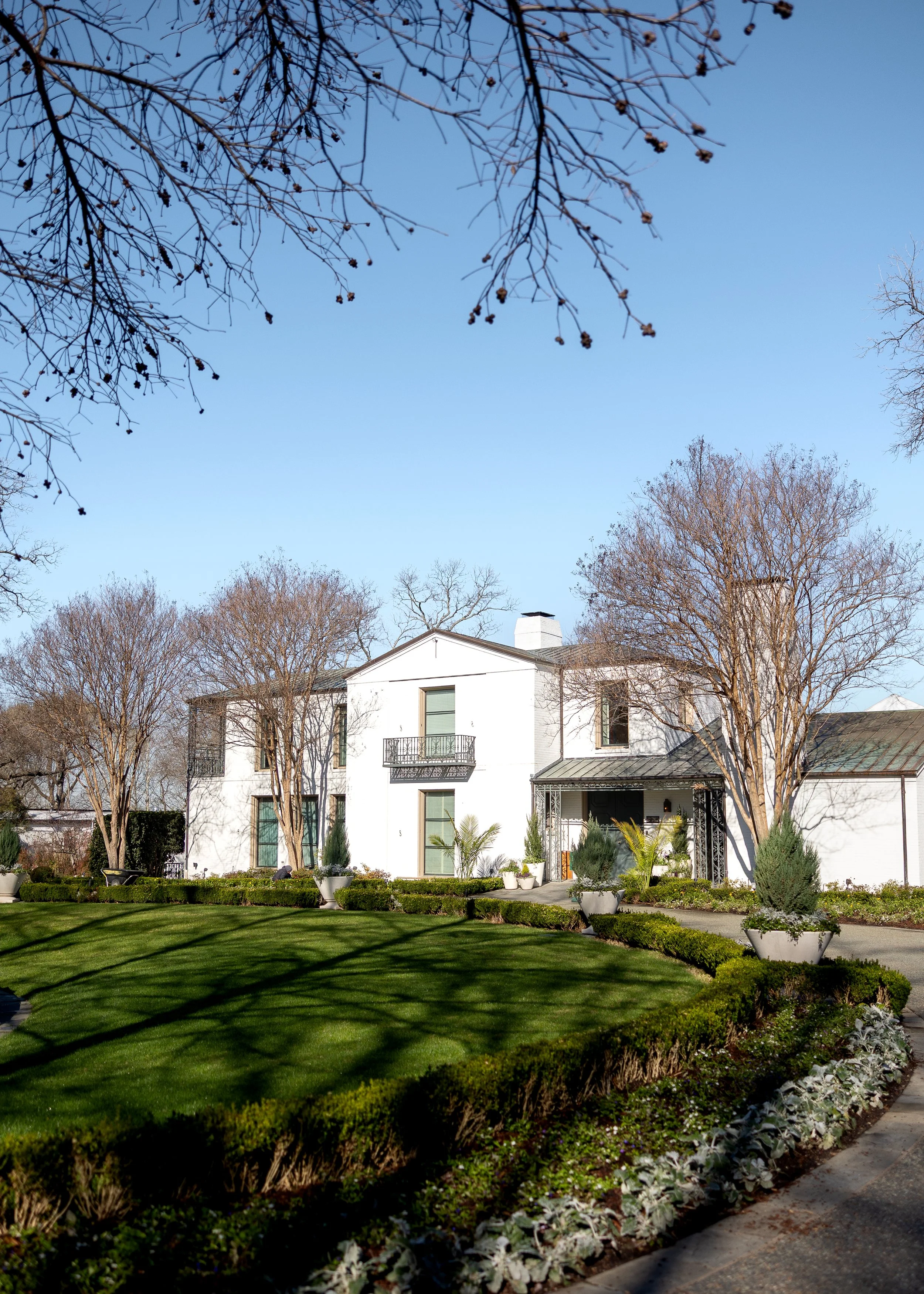 A large white two-story house with a porch, surrounded by a well-manicured garden with shrubs and trees, under a clear blue sky. Camp House, Dallas Aroboretum
