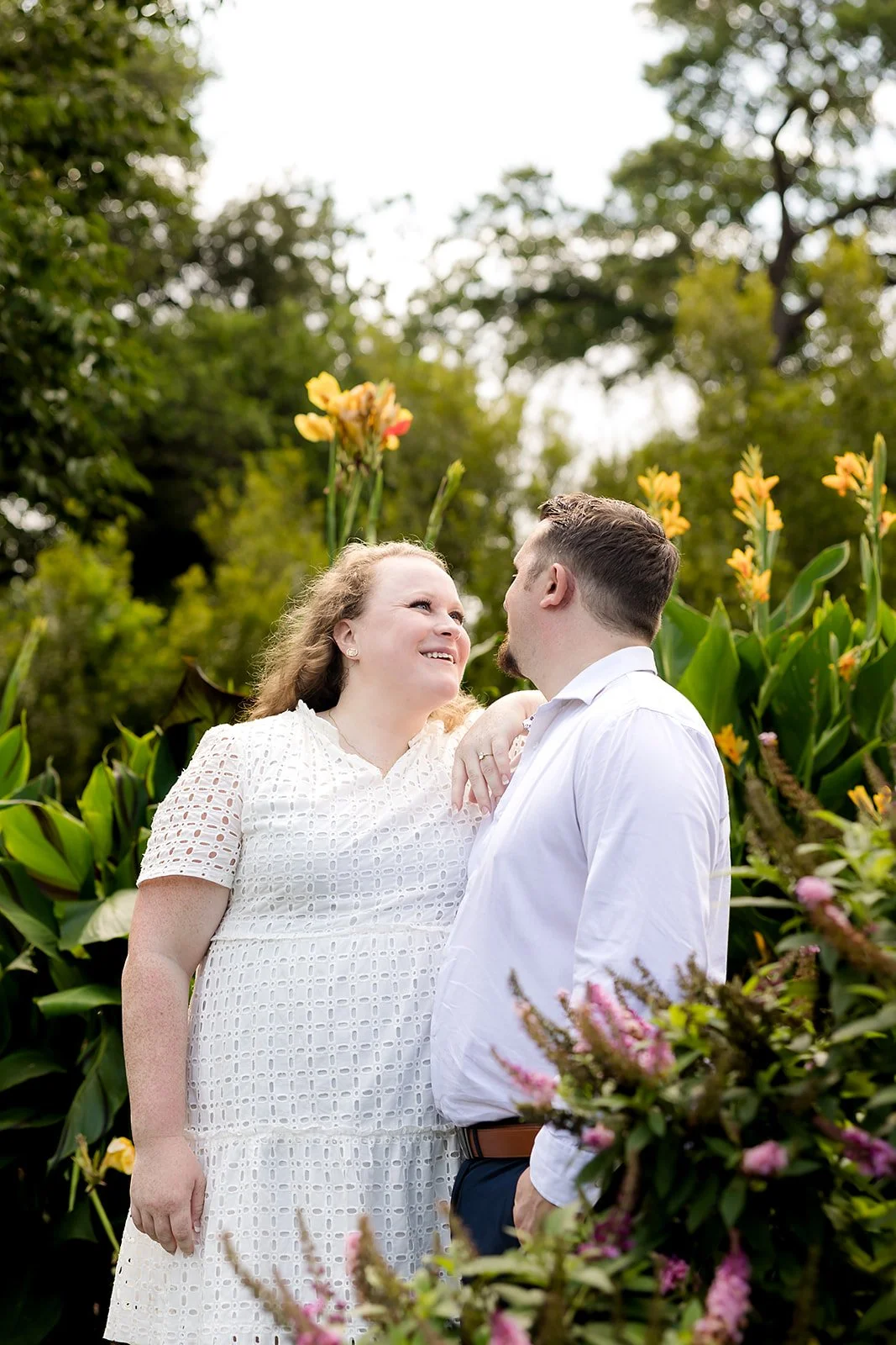 A couple standing outdoors in a garden with yellow and pink flowers, looking at each other and smiling.