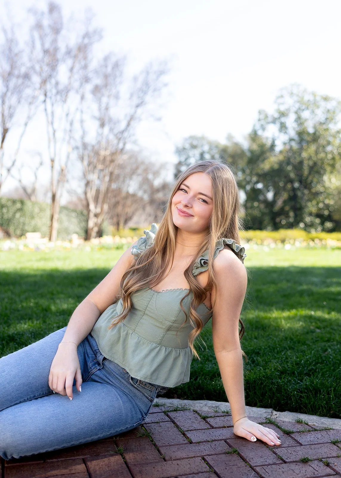 Young woman with long wavy hair sitting outdoors on a brick pathway, smiling and looking at the camera, with green grass and trees in the background.