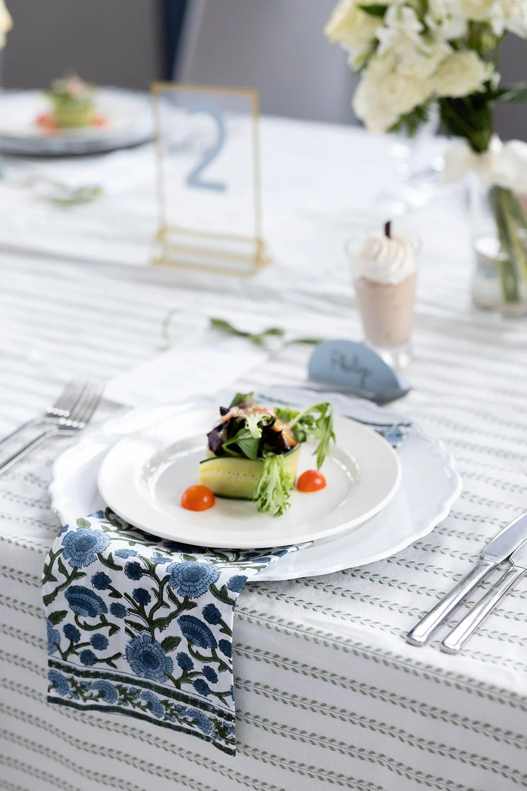 Elegant table setting with a plate of salad and cherry tomatoes, a floral napkin, cutlery, a cupcake, and a flower arrangement in the background.