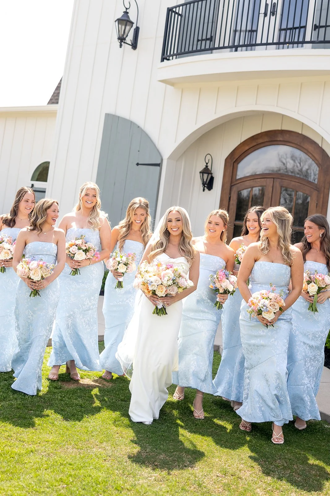 Bridal party of nine women in pastel blue dresses with bouquets, standing outside a white building with wooden door, on a sunny day.