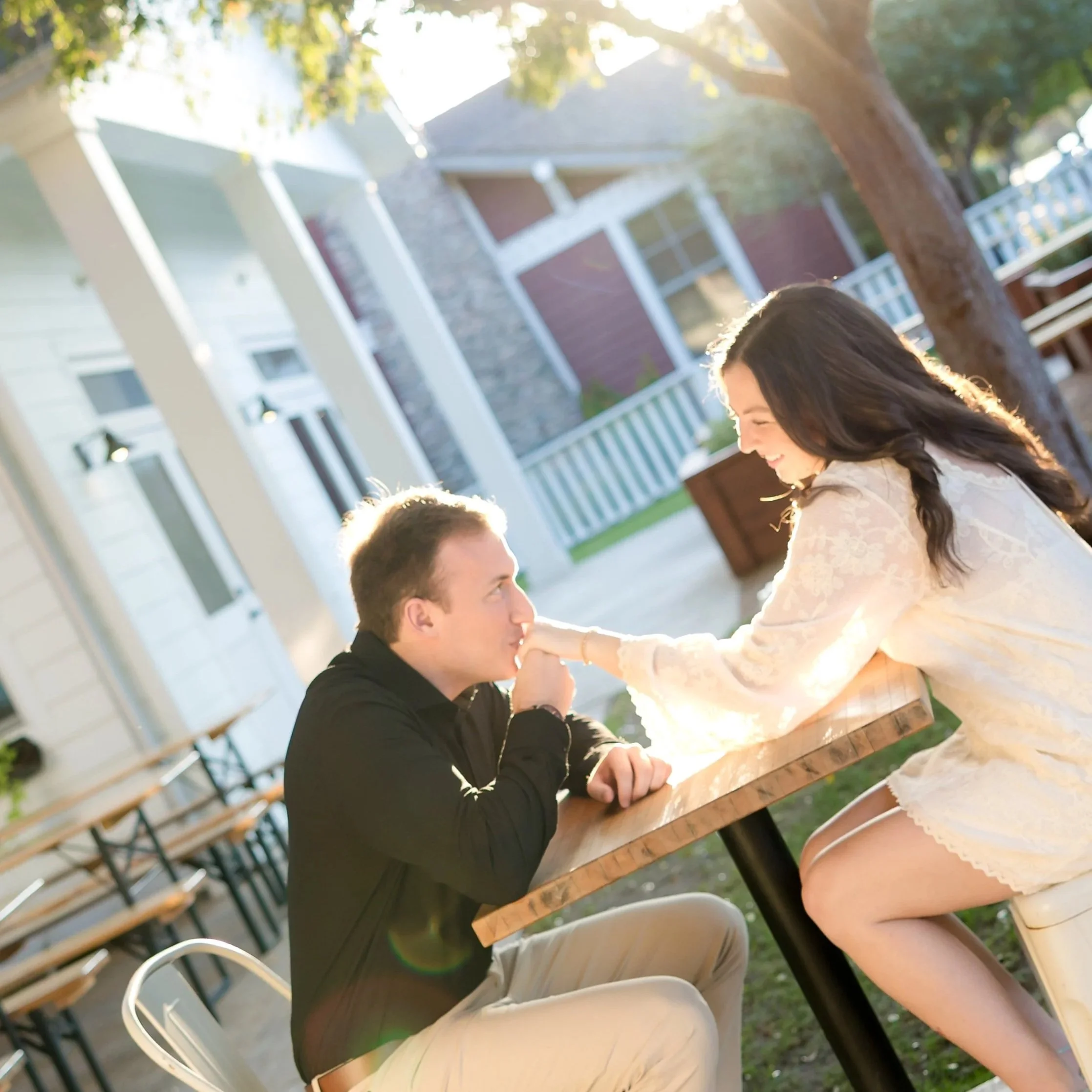 A young woman and man sitting at a wooden outdoor table, smiling and touching noses, with sunlight filtering through trees in the background.