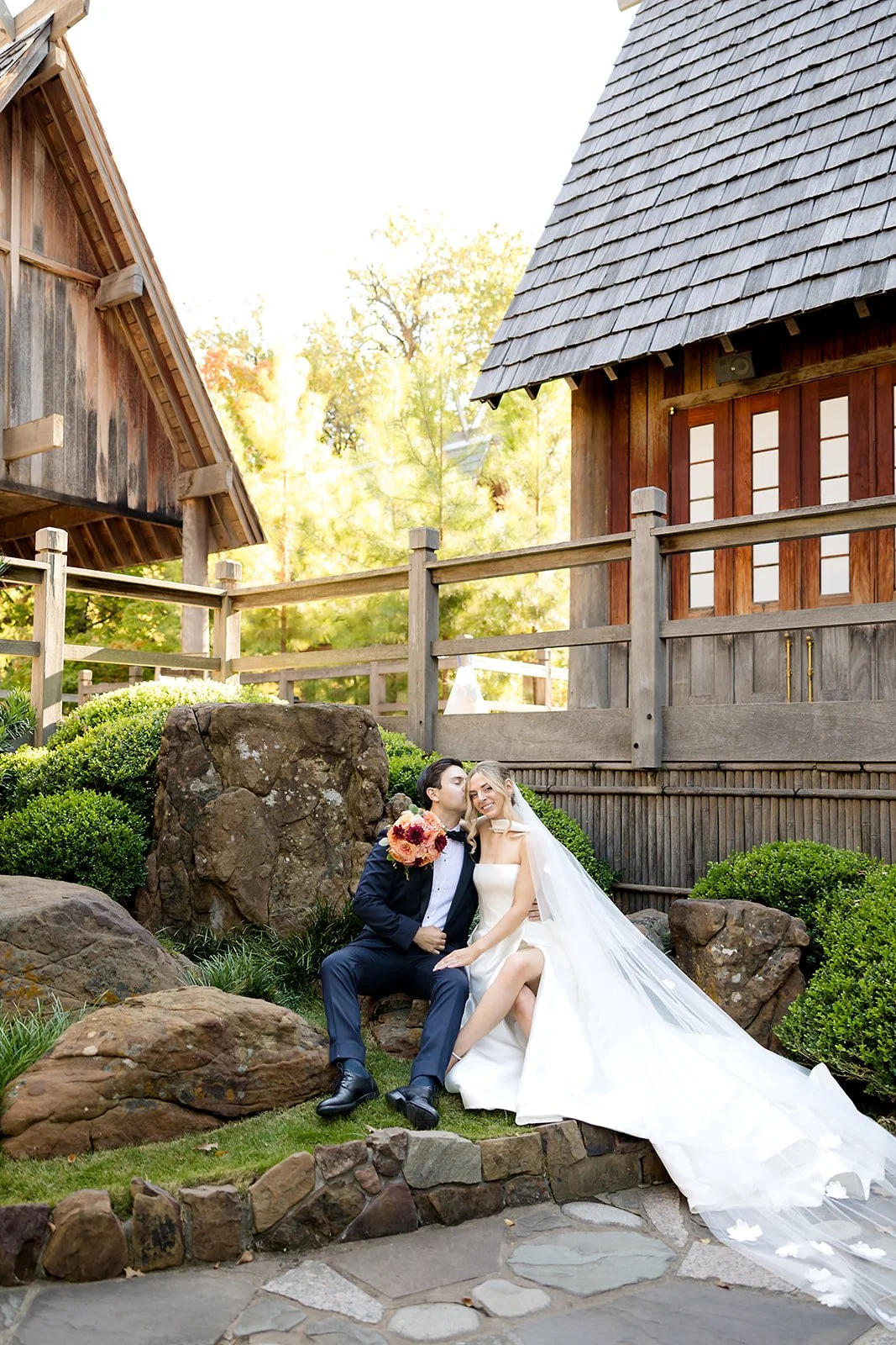 A newlywed couple sitting on rocks outdoors at Fort Worth Botanical Gardens, with a rustic wooden fence and buildings in the background, the bride in a white dress with a long veil and the groom in a dark suit, holding a bouquet of flowers.