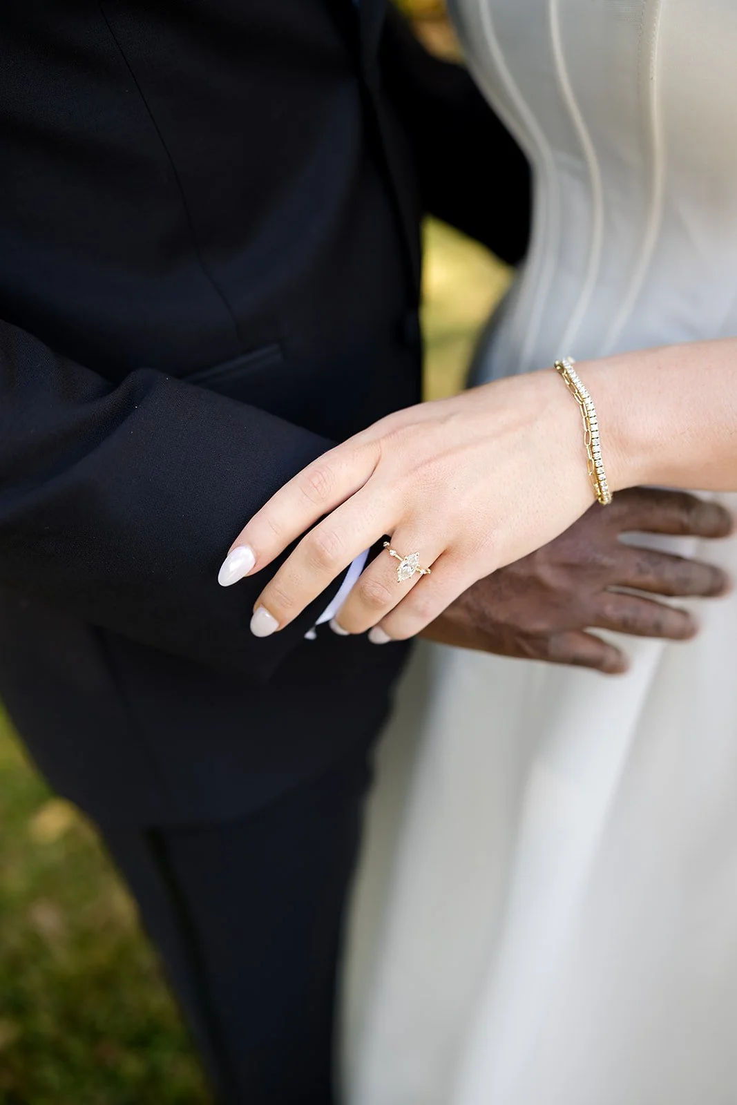 Close-up of a newlywed couple's hands with wedding rings; the bride's hand rests on the groom's arm, showcasing a diamond ring and gold bracelet, while the groom's hand is on her waist, with a black suit and a white dress.