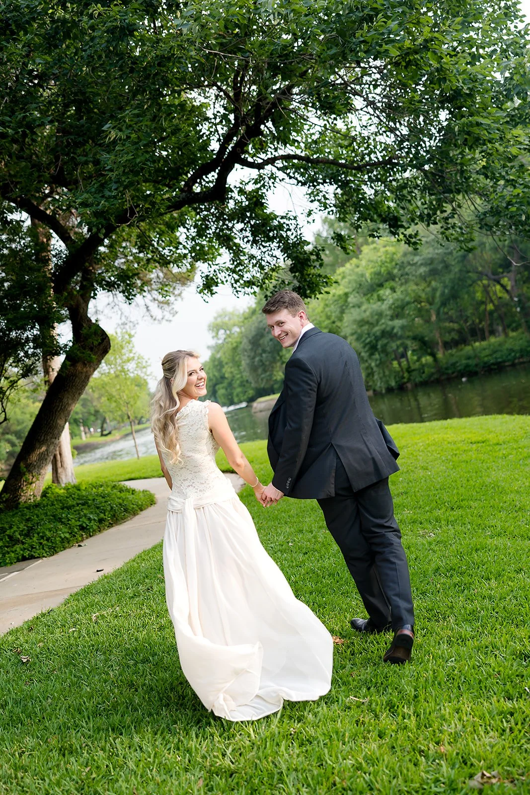A happy newlywed couple holding hands and smiling, walking through a park with green grass, trees, a pond, and a sidewalk on a clear day.