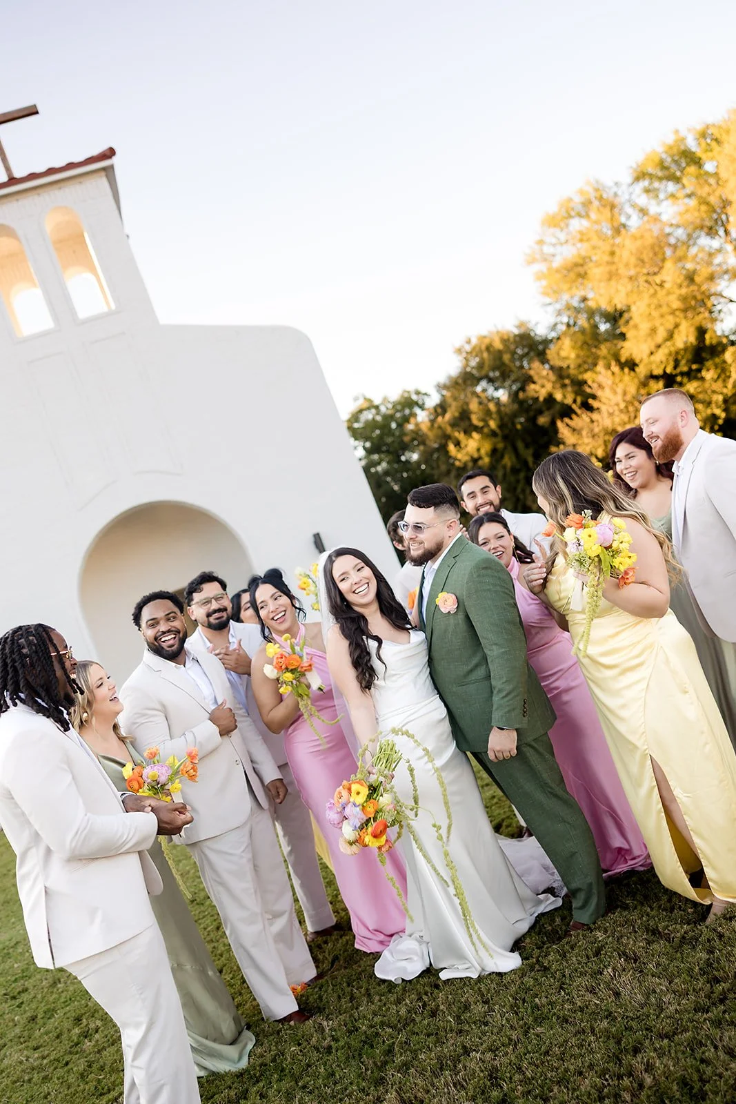 A wedding party stands outside a white chapel, with the bride and groom in the center smiling, surrounded by bridesmaids and groomsmen holding colorful bouquets, during sunset.