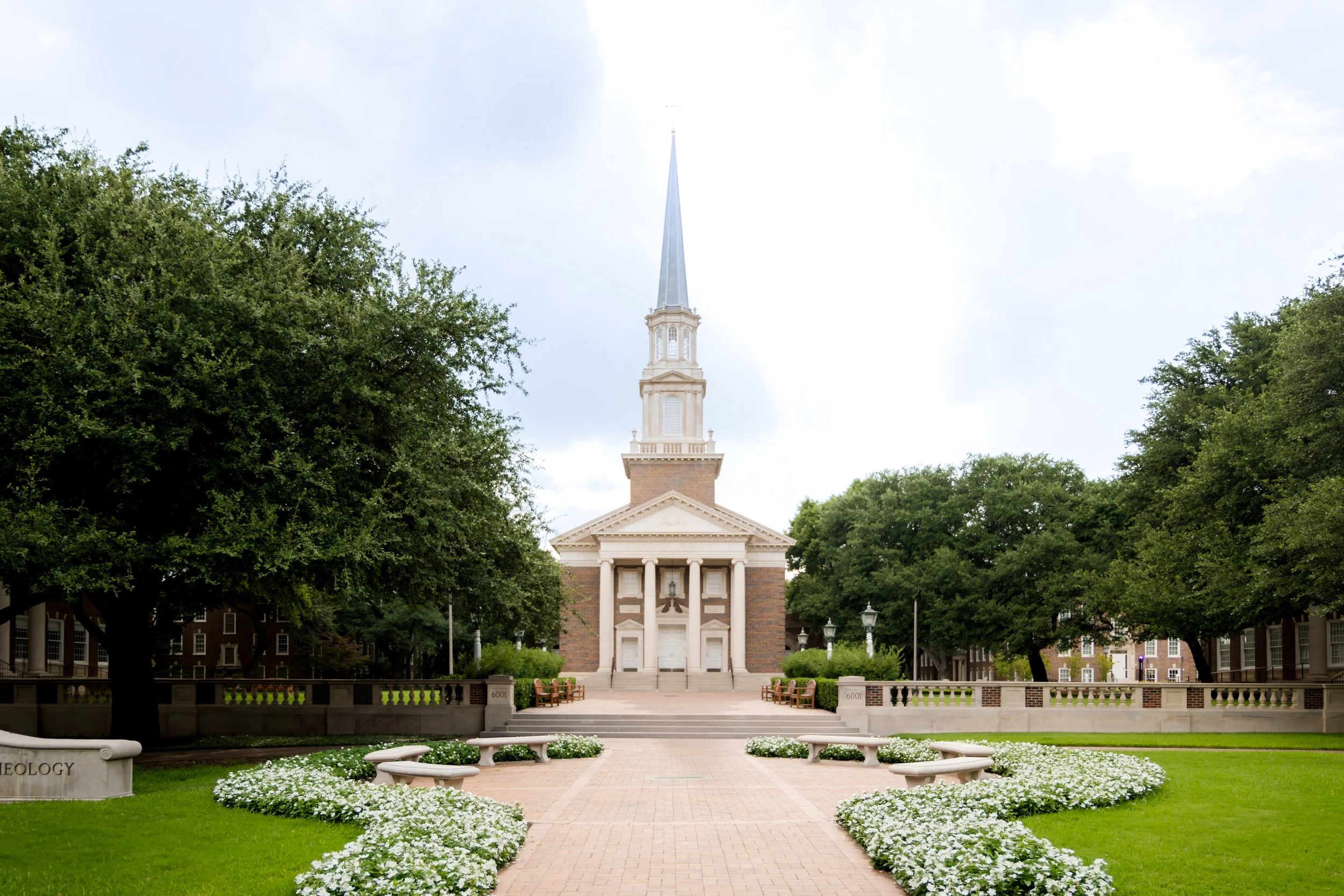 A church with a steeple surrounded by trees and a well-maintained garden with white flowers, benches, and brick pathways. SMU Chapel Wedding.