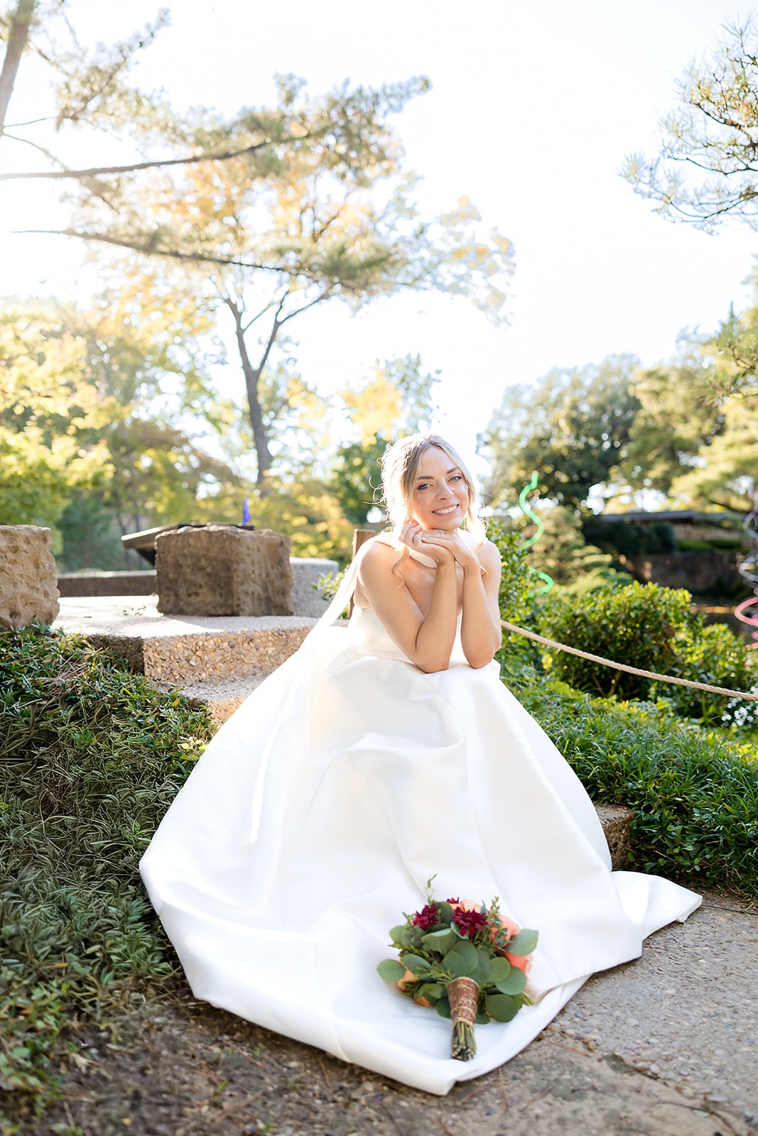 Bridal portrait of a young woman in a white wedding dress, sitting outdoors on a garden path, smiling with hands resting on her chin, small bouquet on the ground in front of her, green trees and bright sky in the background. Dallas Texas