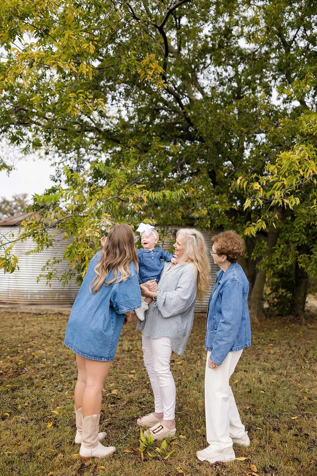 Four women, including one holding a smiling baby girl, are outdoors under a large tree with green leaves. They are all wearing casual clothing, with three women in blue denim and one in a gray and white striped top. The scene appears joyful and relax
