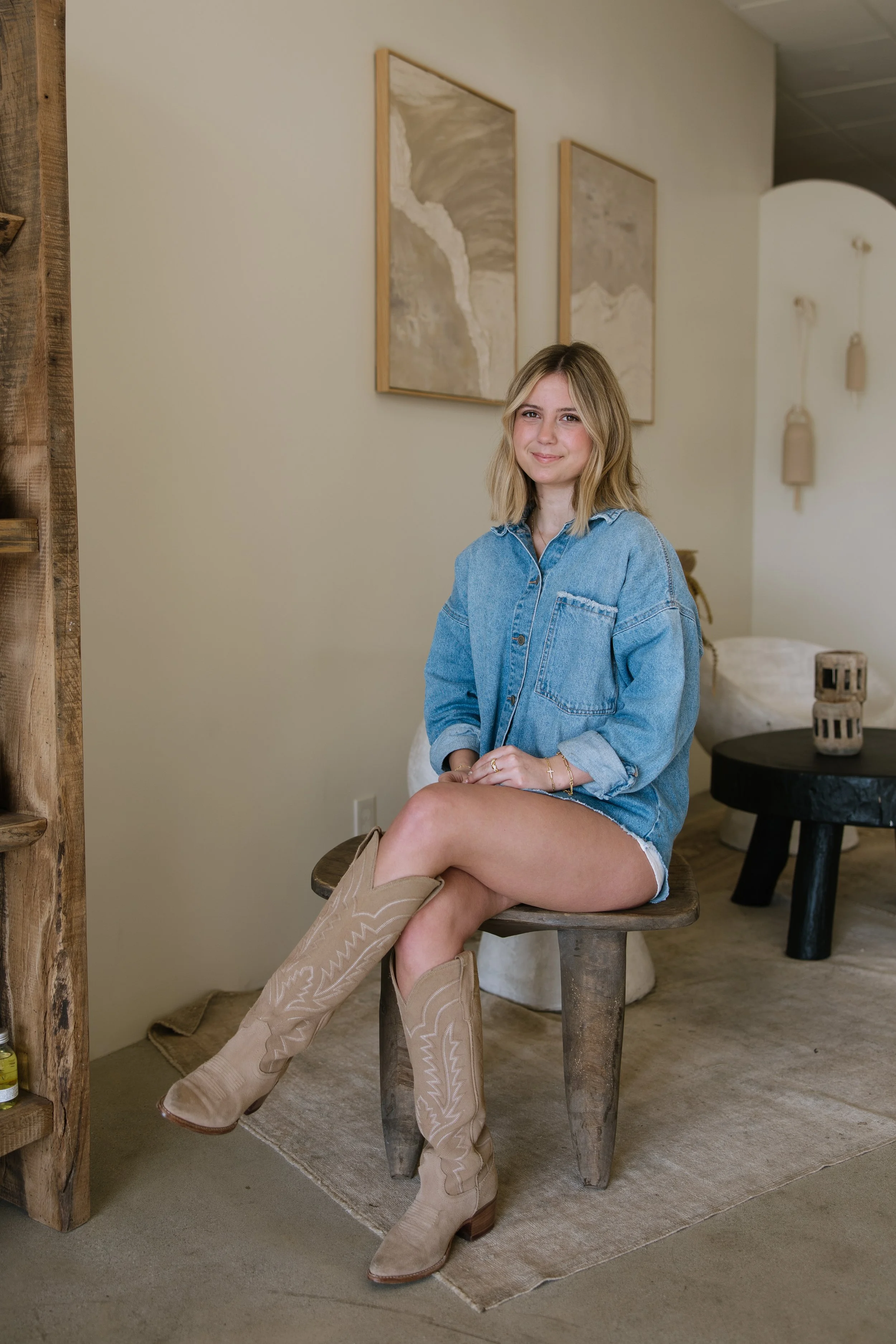 A young woman with blonde hair, wearing a denim shirt and beige cowboy boots, sitting on a wooden stool inside a tastefully decorated room with minimalist art and neutral decor.