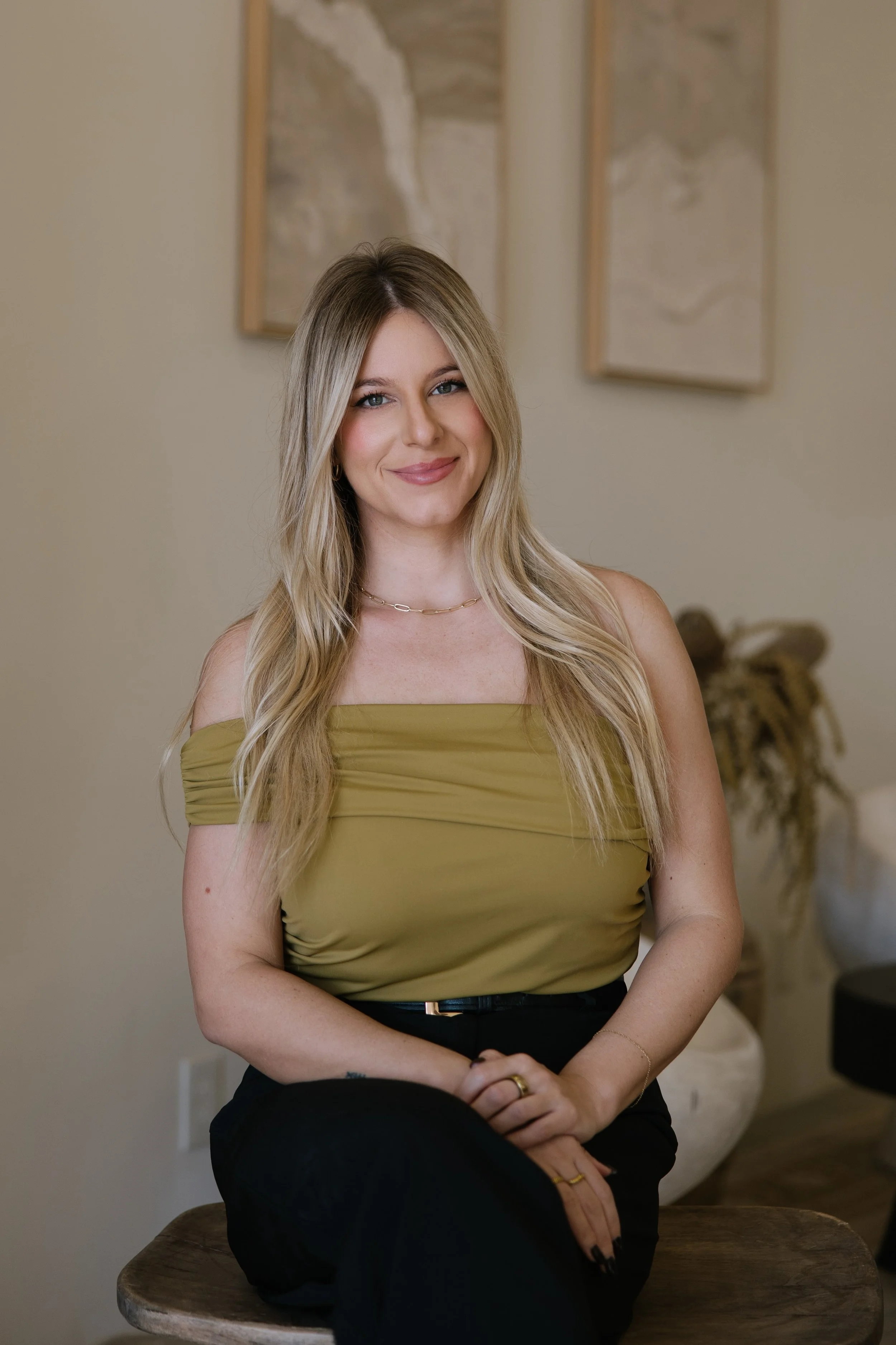 A smiling woman with long blonde hair, wearing an olive green off-the-shoulder top and black pants, sitting on a wooden stool in a room with beige walls and abstract wall art.