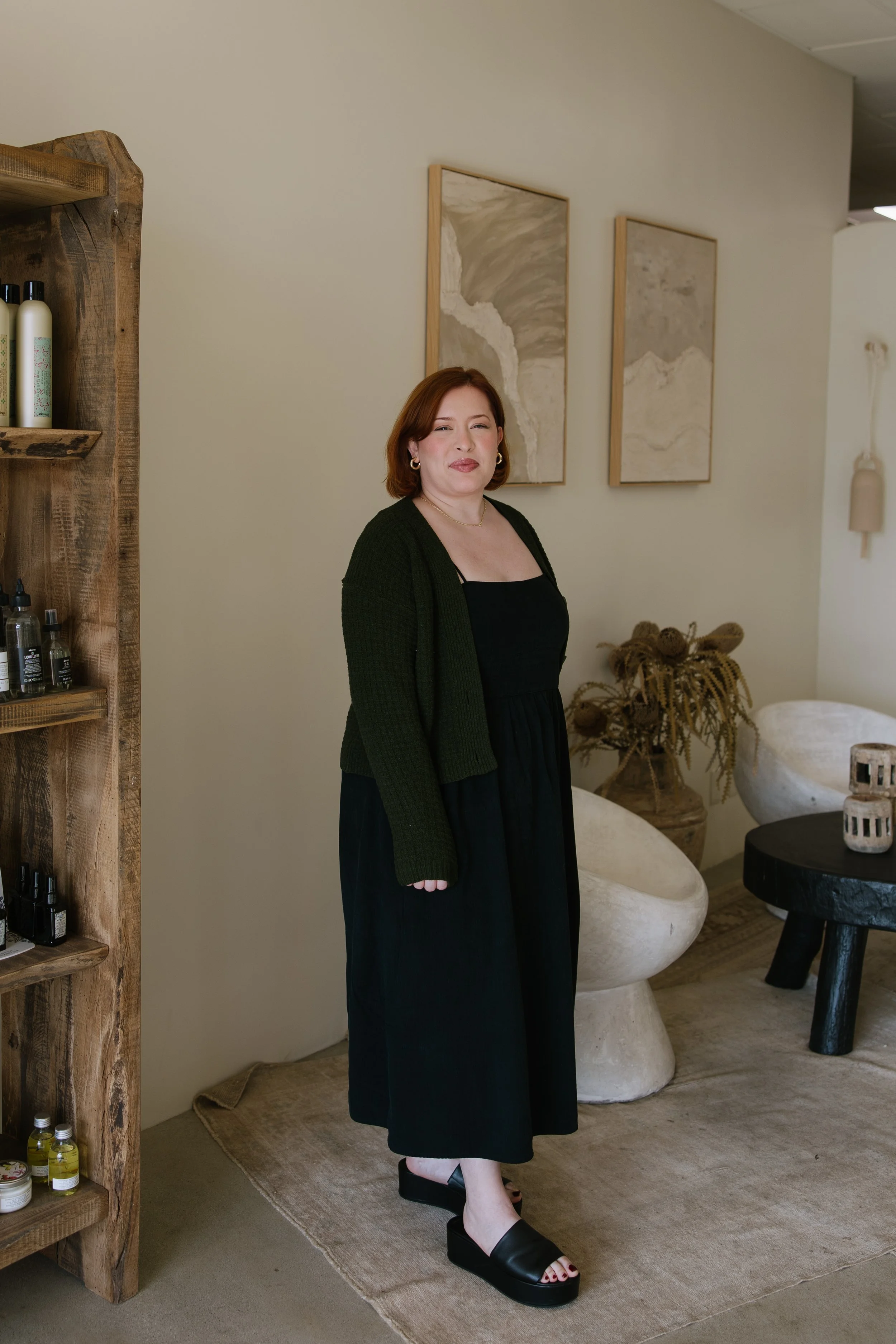 A woman standing in a modern, minimalistic room with beige walls and abstract artwork. She has short red hair, wears a black dress with a black cardigan, and black platform sandals.