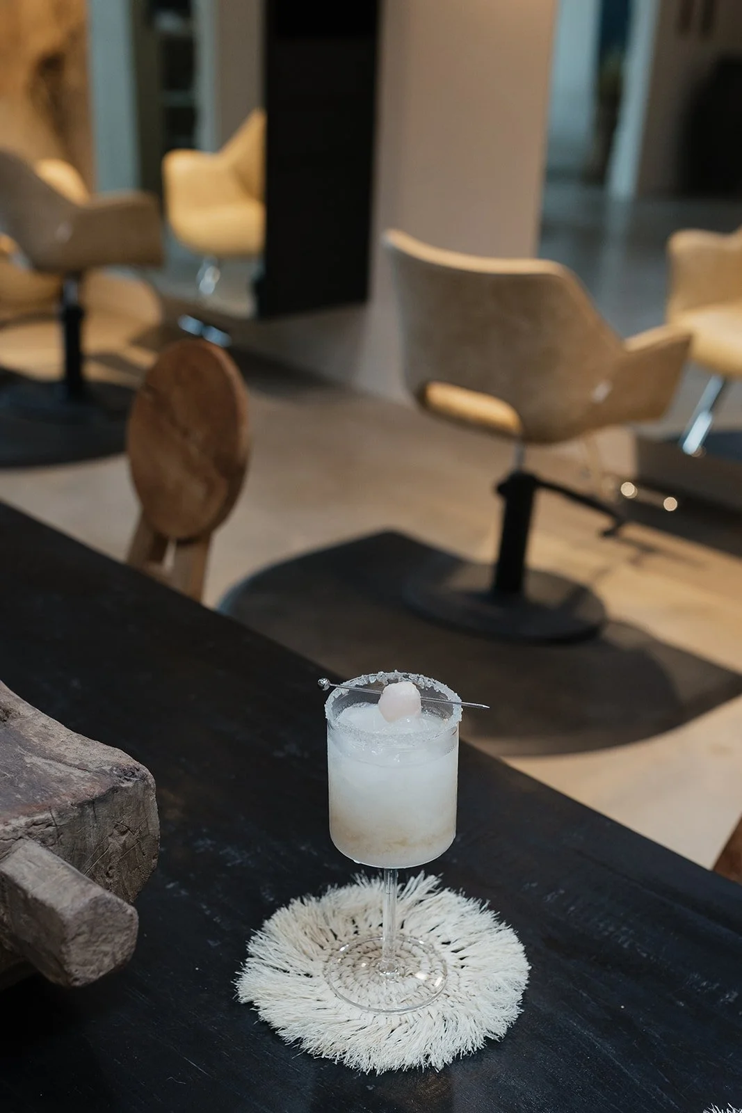 A white cocktail with a cherry garnish in a stemmed glass on a white doily, placed on a dark wooden table in a modern bar or lounge with beige swivel chairs in the background.