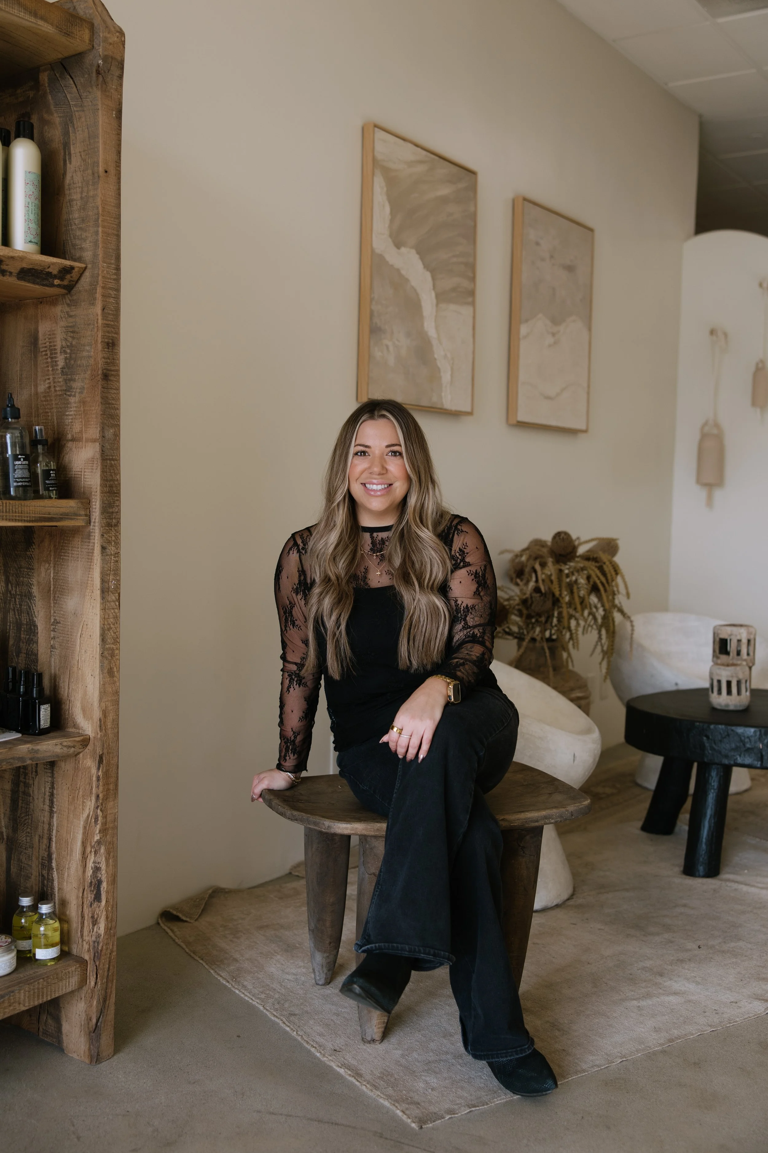 A smiling woman with long wavy hair, wearing a black lace top and black pants, sitting on a wooden stool in a cozy, modern room with earthy decor and abstract artwork on the wall.