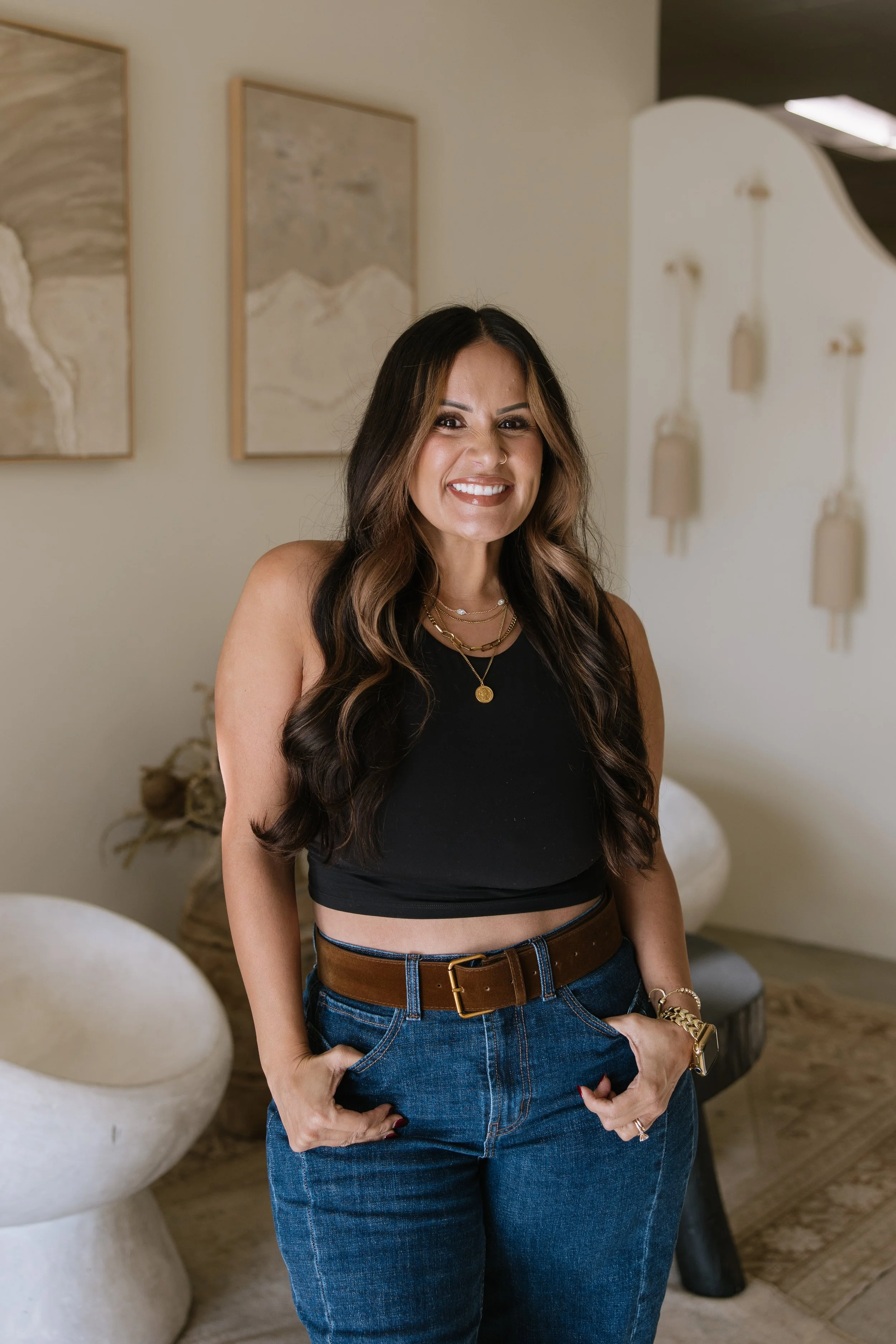 A woman with long, wavy brown hair smiling and standing indoors.