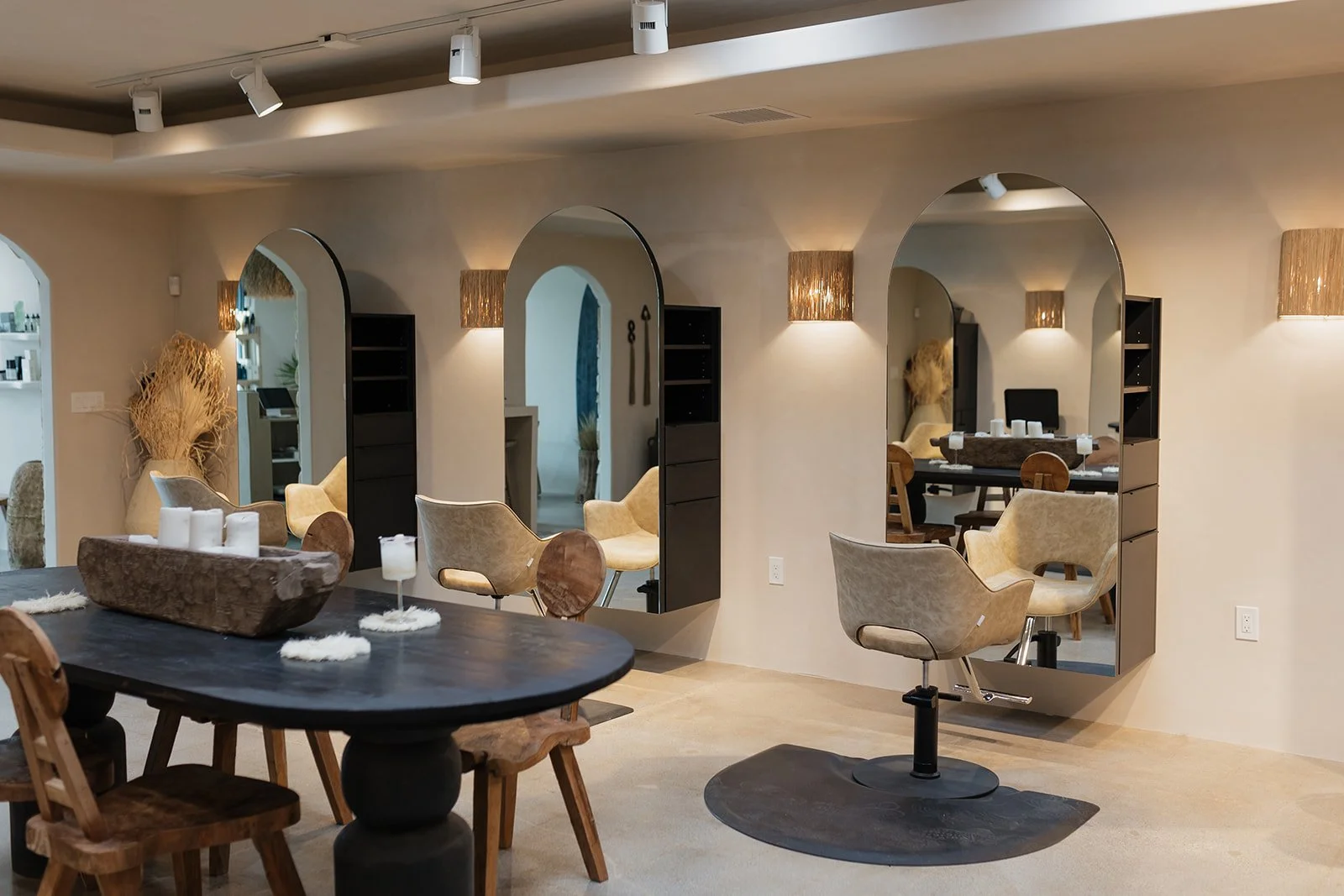 Modern stylistic hair salon with beige walls and multiple large mirrors, including a black shelving unit beside each mirror, beige chairs, and warm wall sconces. The space features a black countertop with white decorative candles, a rustic centerpiece, and a wood dining table with chairs.