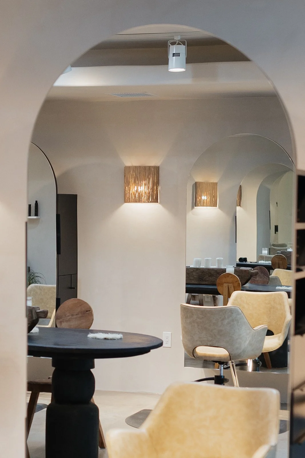 Interior of a modern, minimalist restaurant or cafe with arches, light-colored walls, wooden wall sconces, and chairs in beige and yellow around dark tables.