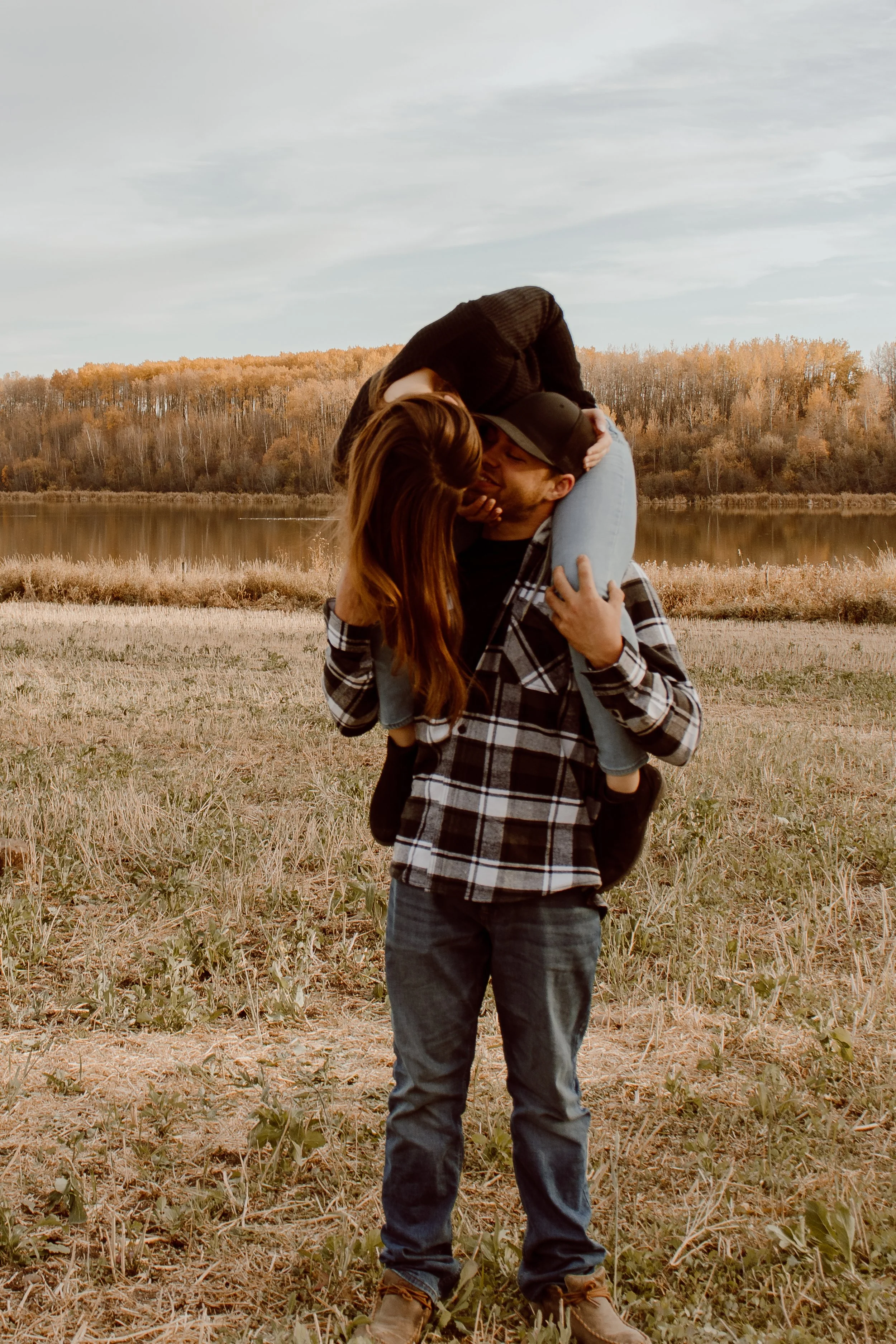 A couple is near a lake with trees in the background. The man is holding the woman on his shoulders, and they are kissing.