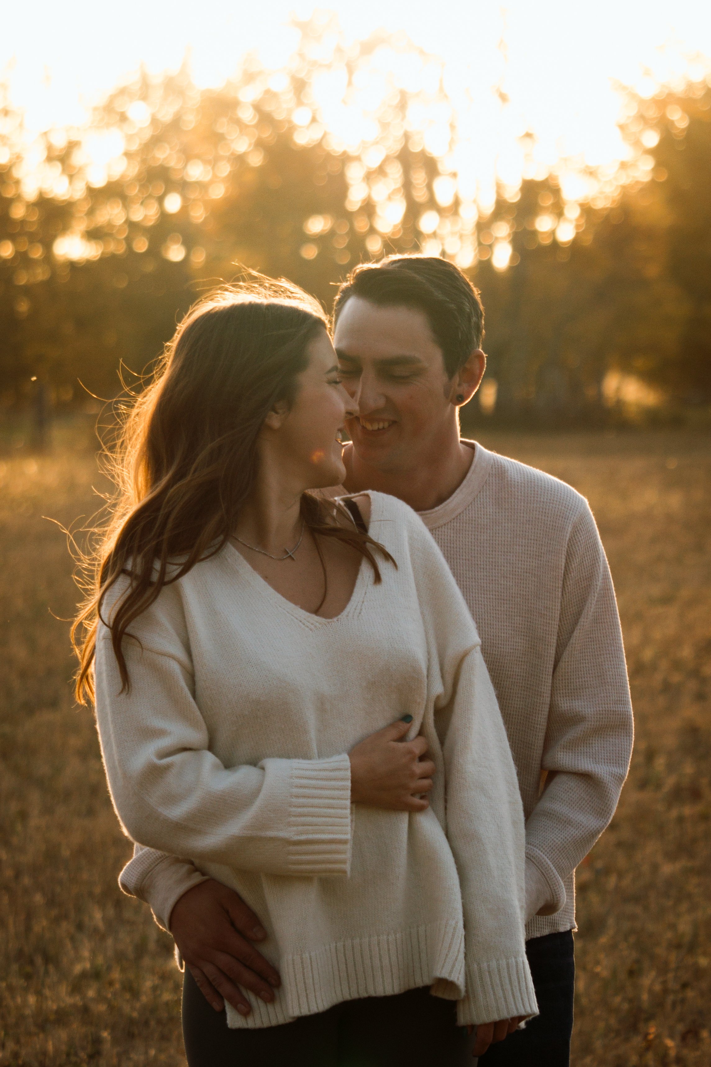 A couple standing close together in a field at sunset, sharing a tender moment with smiling faces.