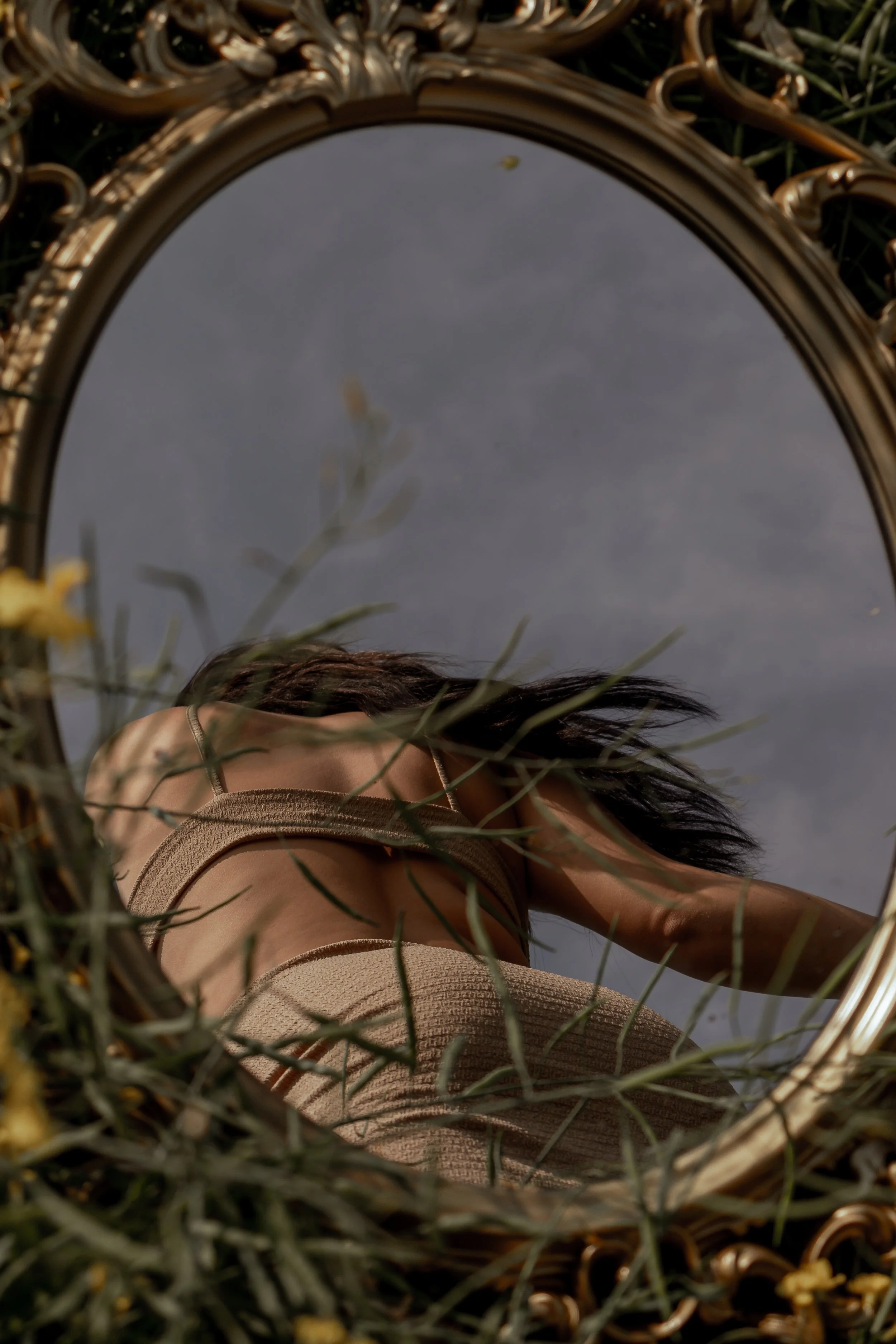 A person with long dark hair is looking into a decorative mirror with a gold ornate frame, placed outdoors on grass, with a cloudy sky reflected in the mirror.