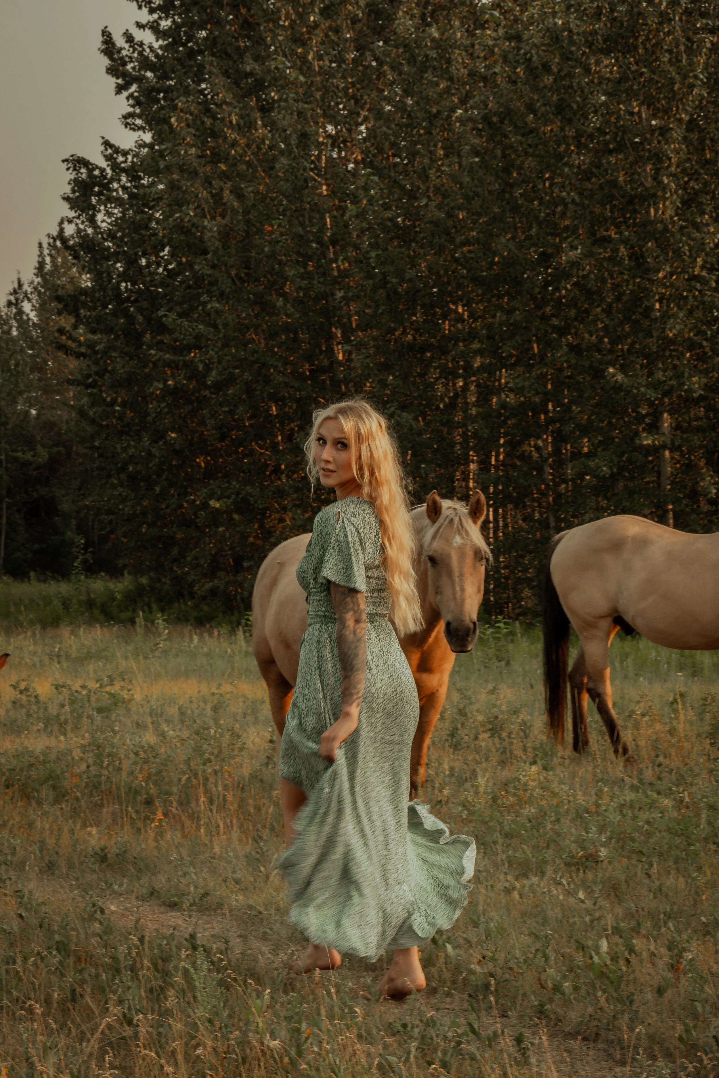 A woman in a long green dress standing barefoot in a field at sunset with three horses, with trees in the background.