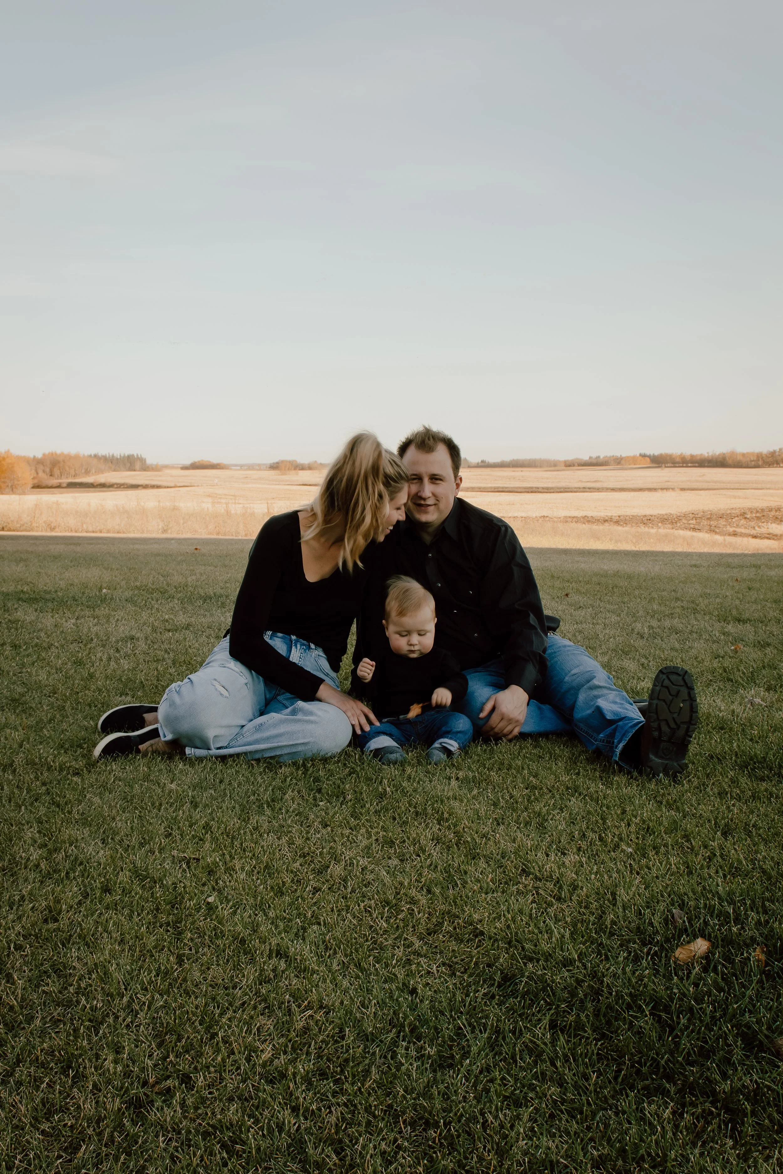 A family of three sitting on grass in a rural field with open farmland and trees in the background, under a clear sky.