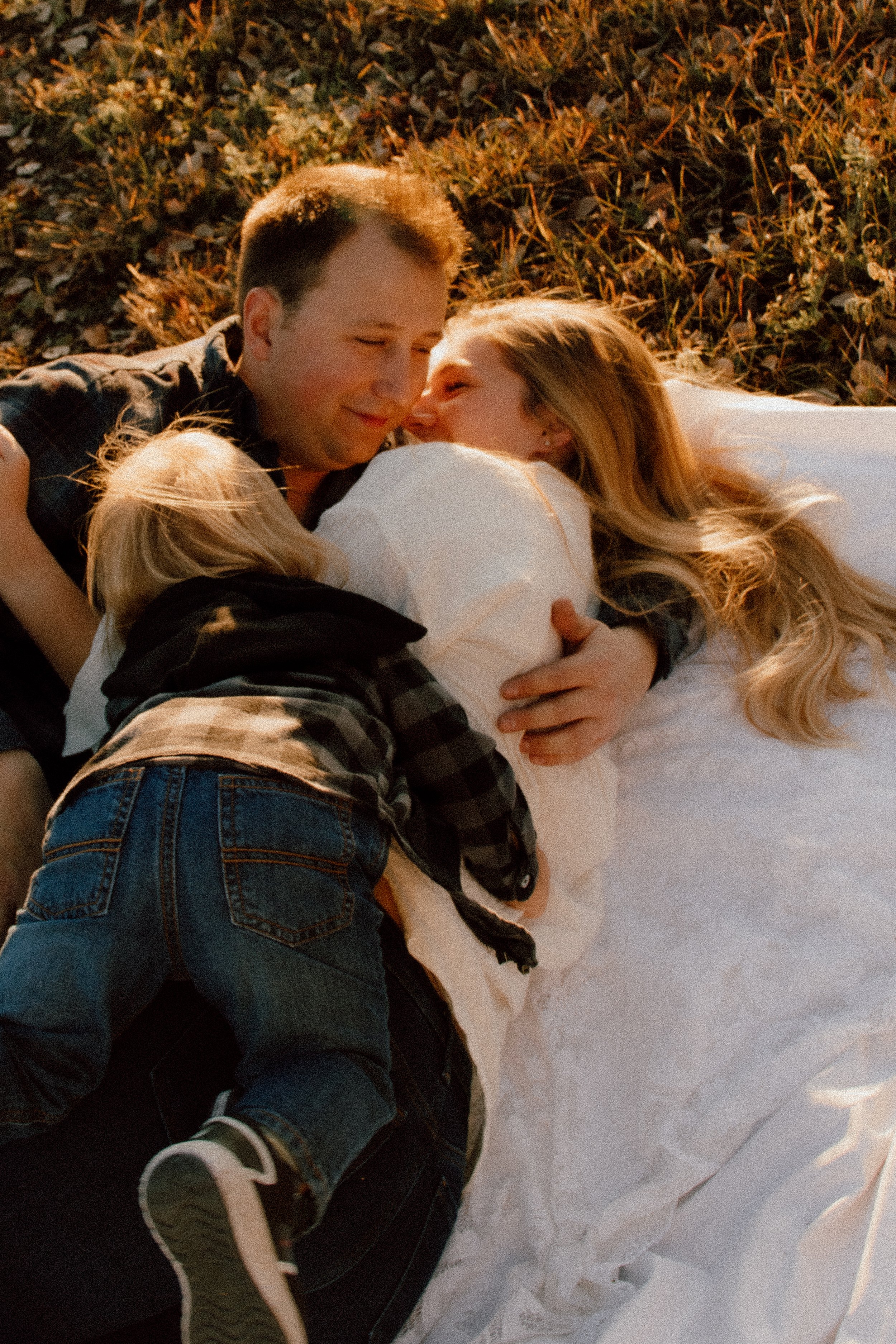 A couple lying on the grass, smiling and cuddling, with a blanket underneath them, during sunset.