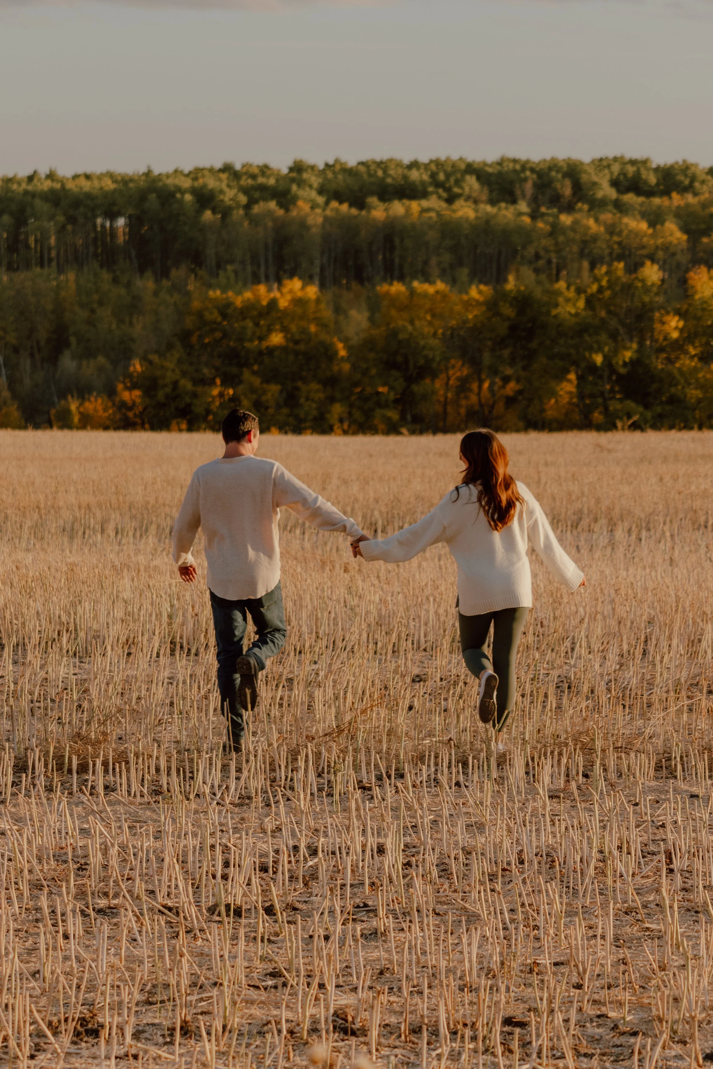 A couple holding hands and running through a harvested field during sunset, with a forest in the background.