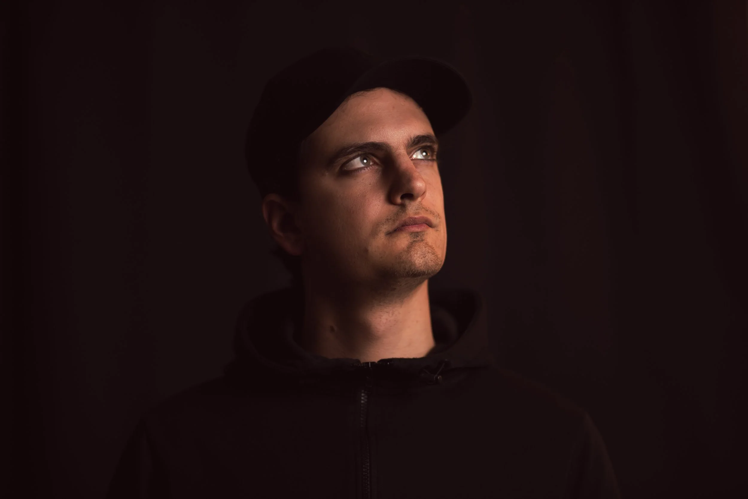 Ritratto di un giovane uomo con cappello nero, vestito con felpa nera, che guarda leggermente verso l'alto, contro sfondo nero.