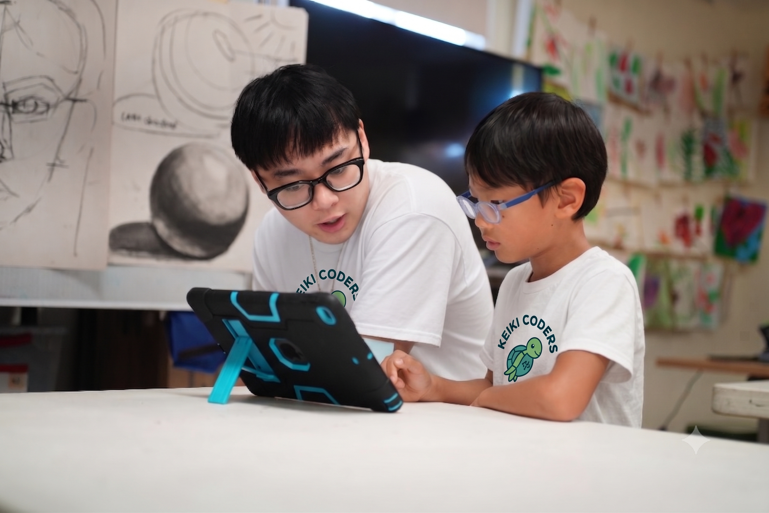 Two boys wearing glasses and white T-shirts with a turtle logo are sitting at a table, focused on a device or project. The background features children's artwork and posters on the walls.