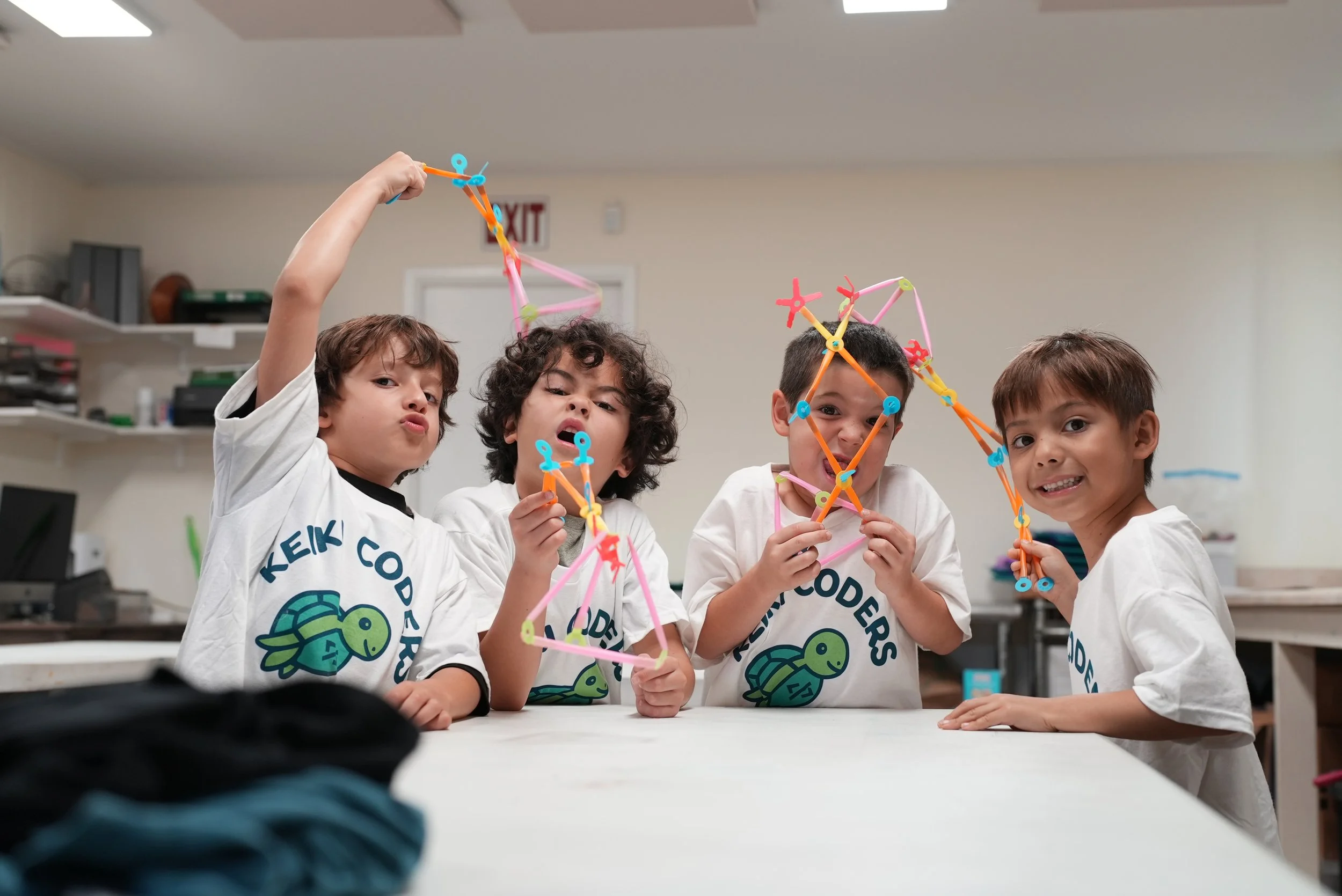 Four children wearing white shirts with turtle logos are holding colorful geometric structures made from connecting sticks and joints, smiling indoors with shelves and an exit sign in the background.