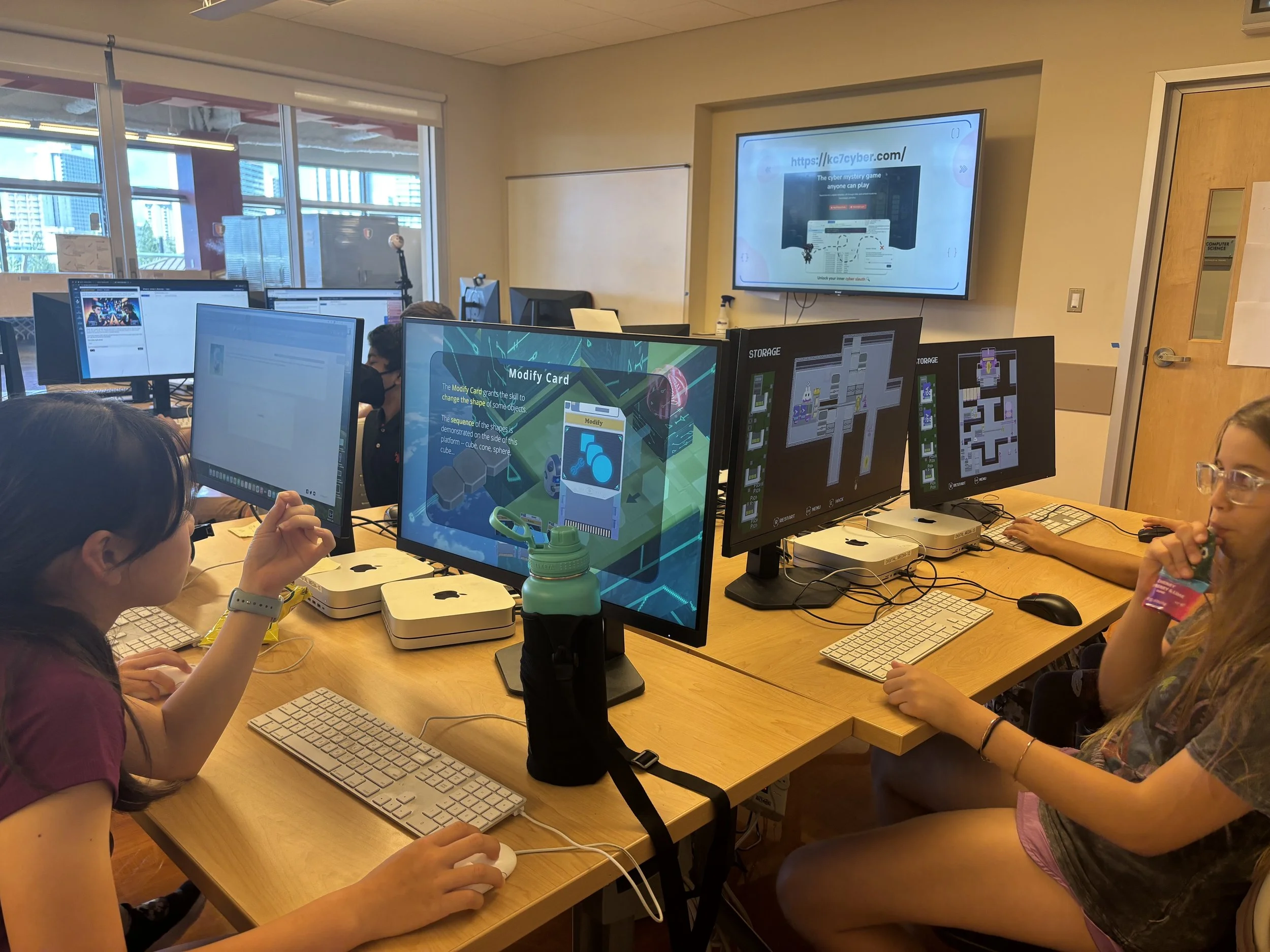 A classroom filled with students working on computers, some looking at screens displaying digital content, with a wall-mounted monitor on the front showing a website.