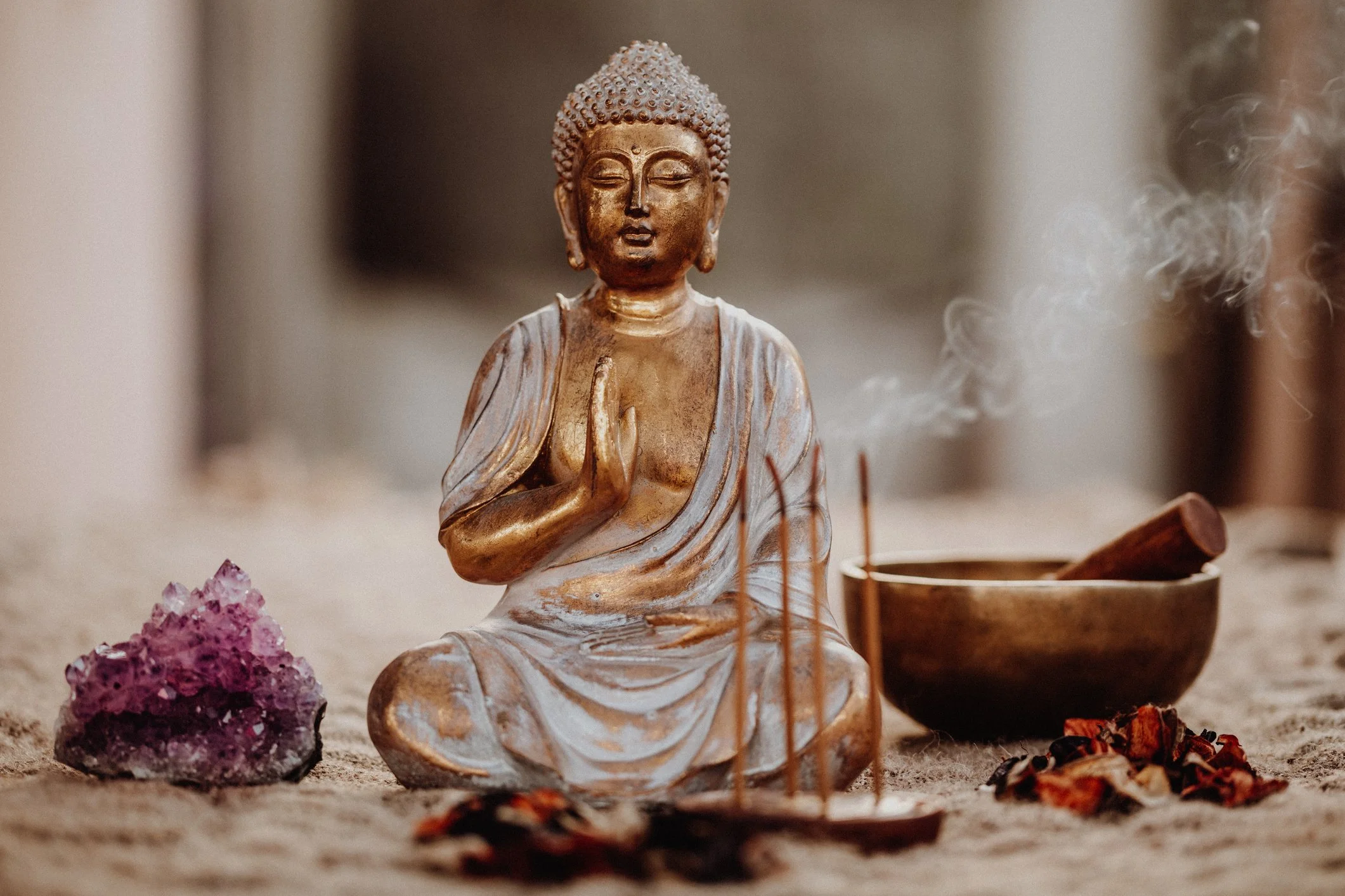 A small Buddha statue with closed eyes and folded hands, surrounded by crystals and spiritual items on a sandy surface.