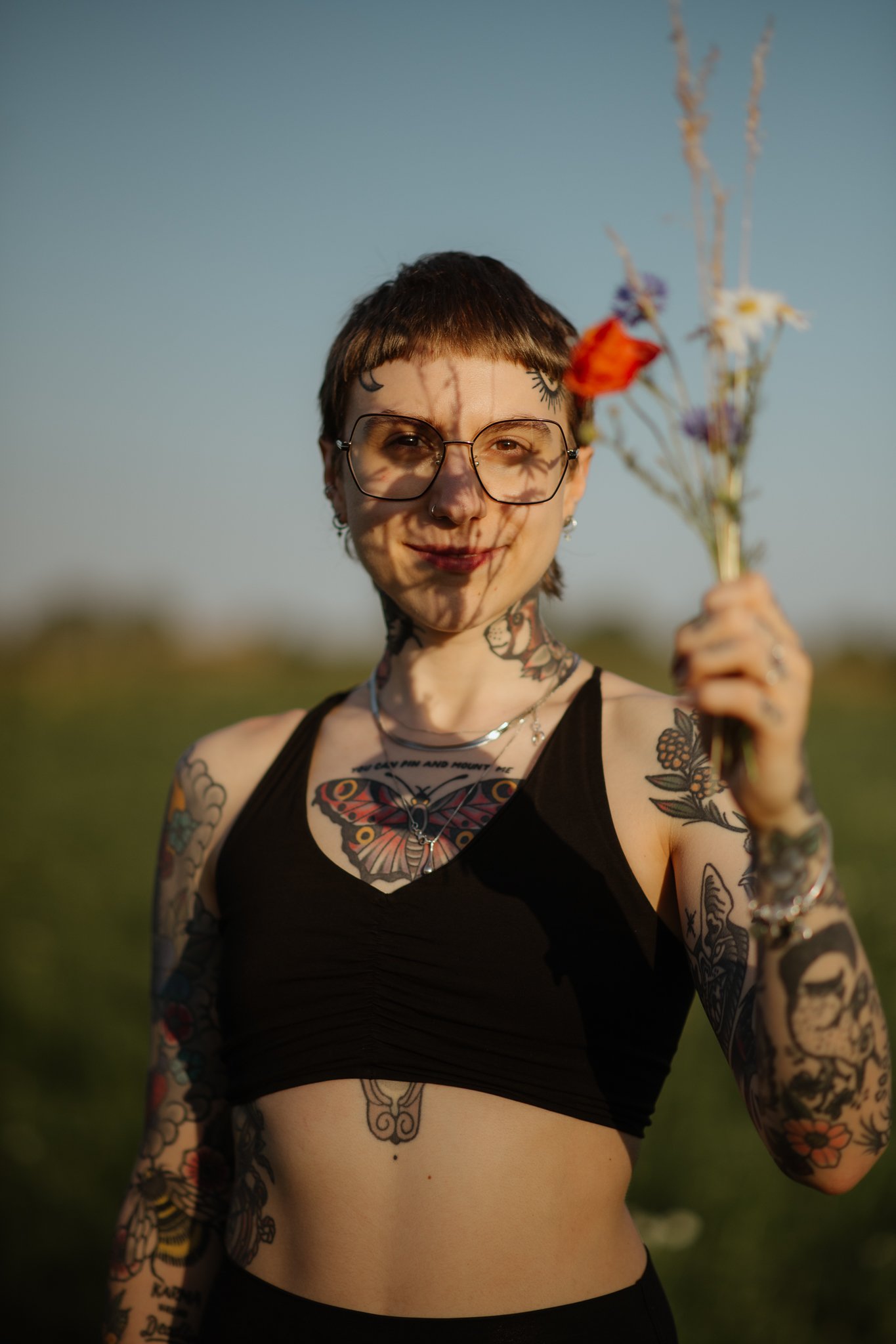 Junge Frau mit Tattoos, trägt Brille, hält einen Blumenstrauß, steht im Freien bei Sonnenuntergang.