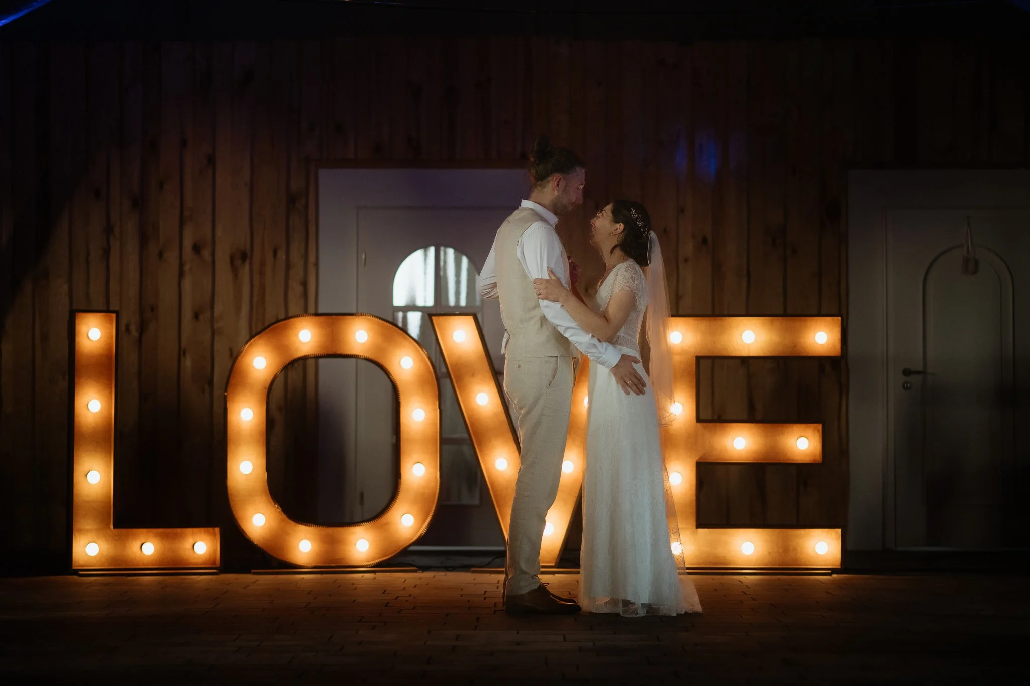 Ein Brautpaar tanzt unter einem großen leuchtenden LOVE-Schriftzug bei einer Hochzeit in einem rustikalen Raum.