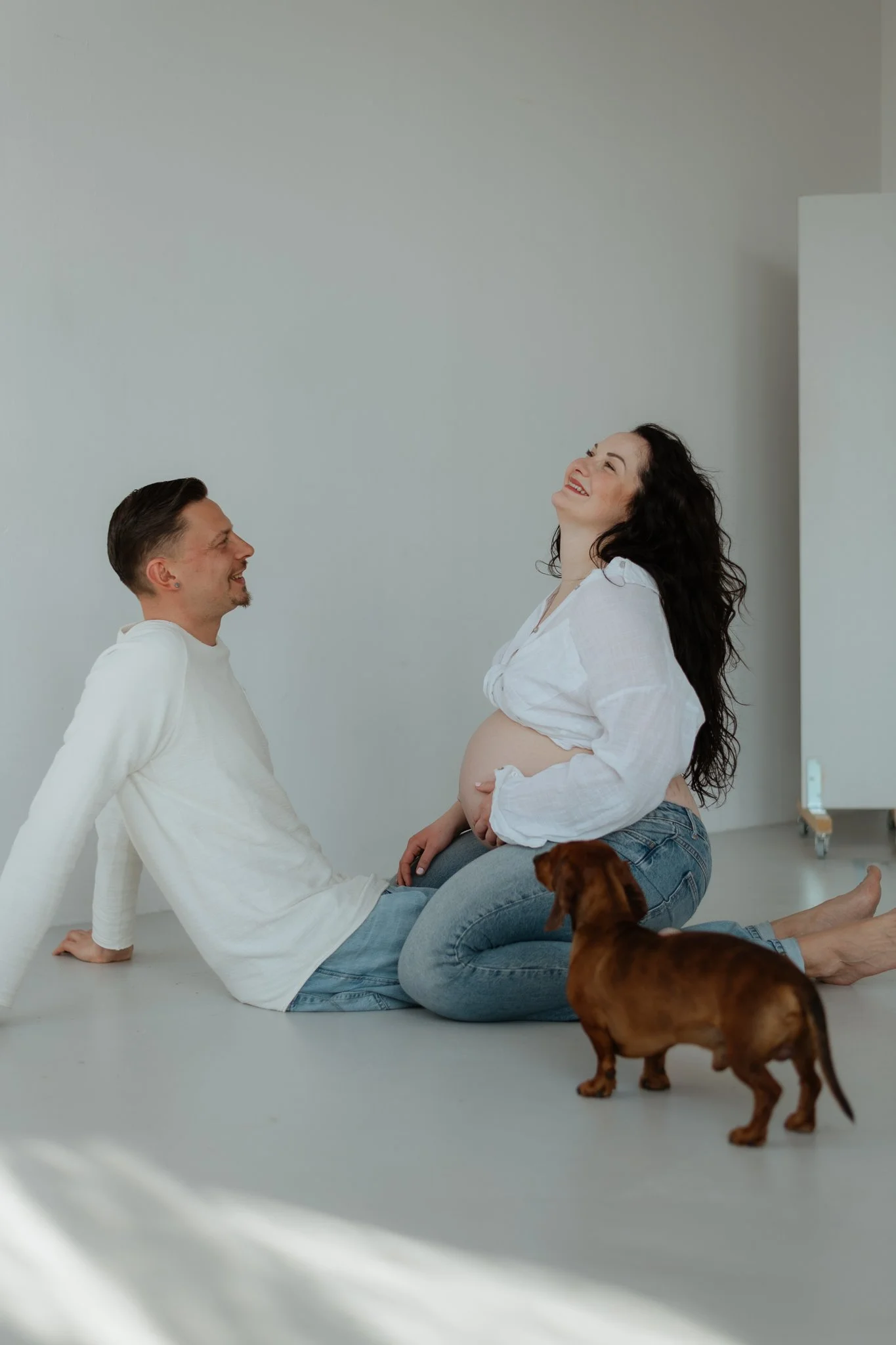 Ein lachendes Paar sitzt auf dem Boden, die schwangere Frau hat dunkle lockige Haare und trägt eine weiße Bluse, der Mann trägt ein weißes Oberteil und blaue Jeans, ein kleines braunes Hundchen steht vor ihnen in einem hellen Raum.