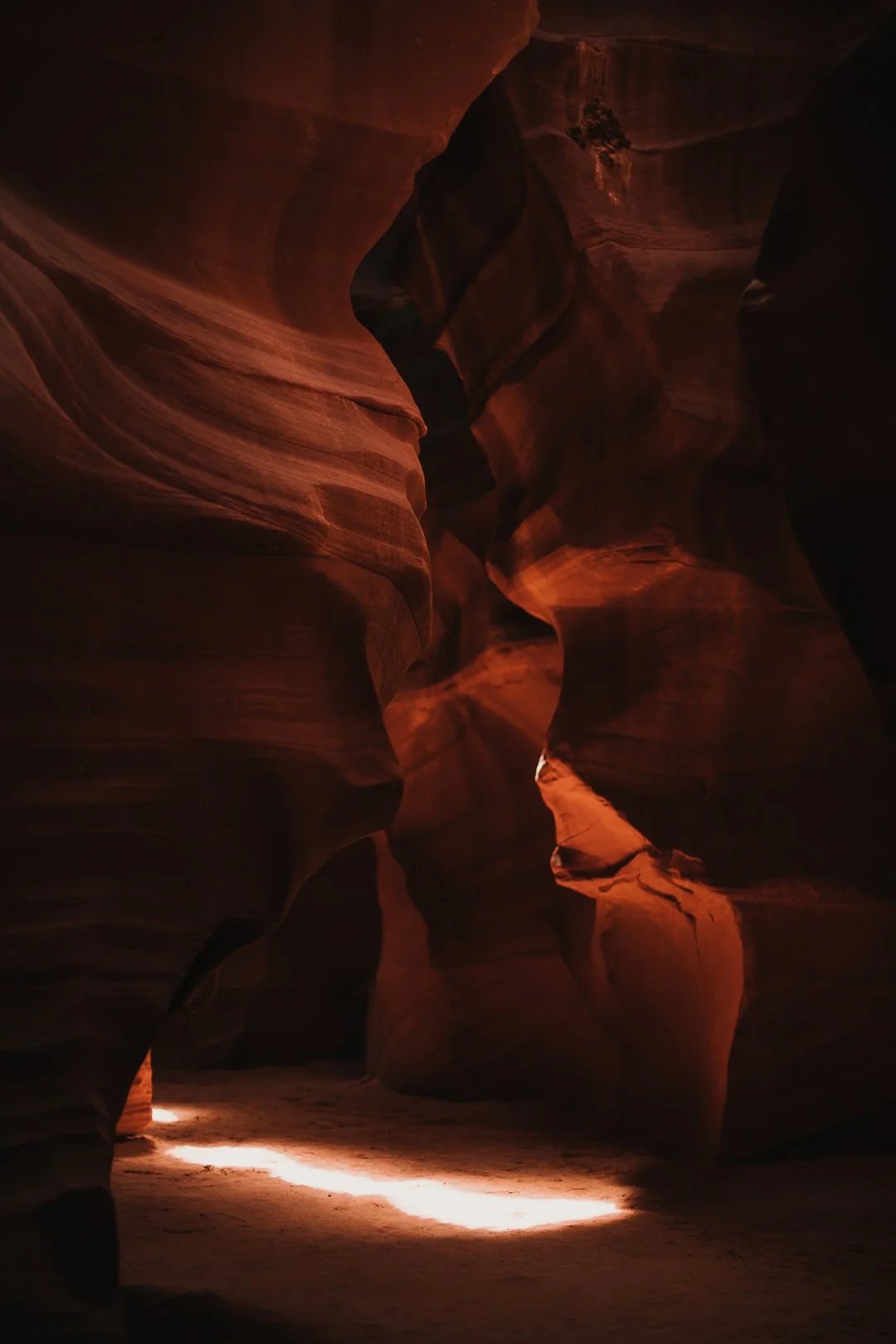 Eine enge, geschwungene Schlucht mit glatt geformten roten Gesteinswänden, durch die Sonnenlicht Streifen auf den Sandboden wirft.