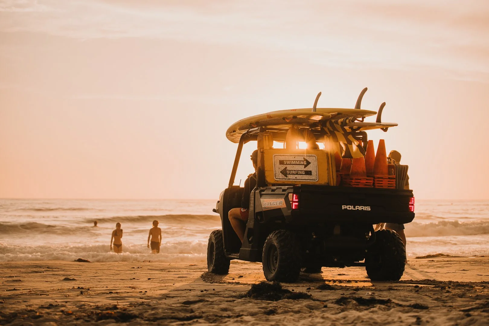 Polaris Geländewagen mit Surfbrett und Verkehrszeichen am Strand bei Sonnenuntergang, im Hintergrund schwimmen Menschen im Meer.