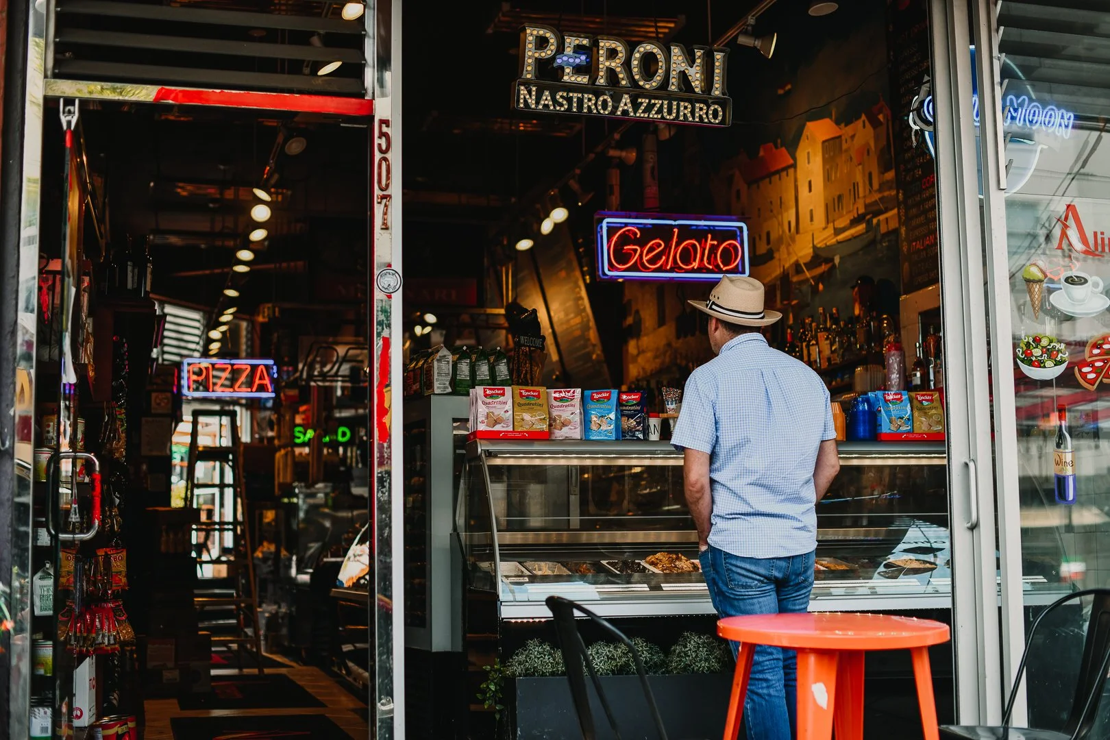 Ein Mann steht vor einer Eisdiele mit neonbeschilderten Hinweisen auf Gelato und Pizza, trägt Hut und blaue Kleidung, im Vordergrund ein roter Tisch.