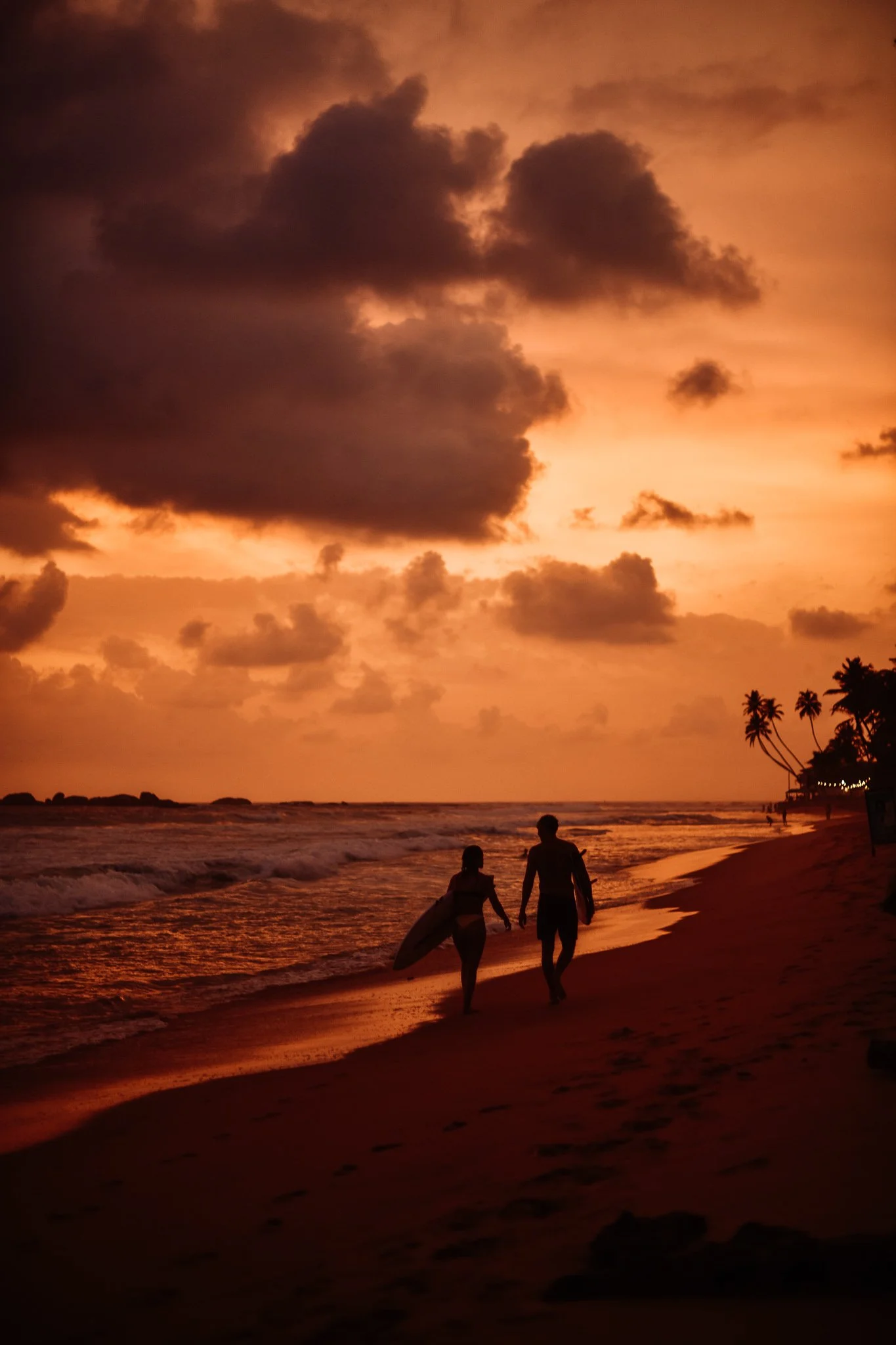 Paar läuft am Strand bei Sonnenuntergang, einer hält ein Surfbrett, Palmen am Horizont.