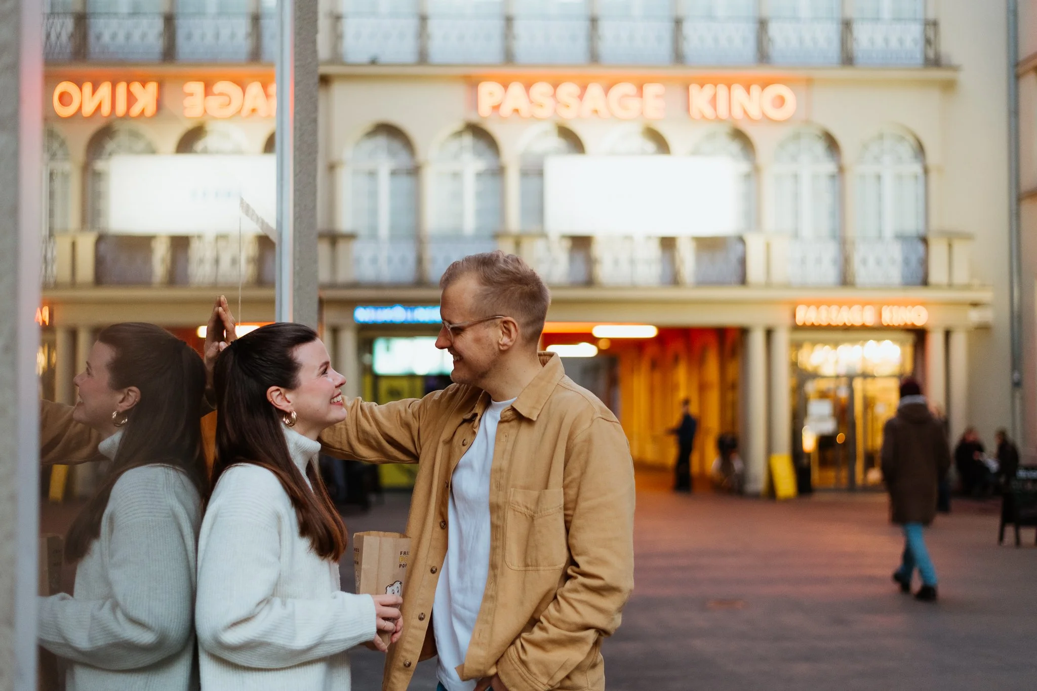 Zwei Frauen und ein Mann lachen sich im Freien an einem Abend an, vor einer Gebäude mit Neonbeschriftung 'Passage Kino'.
