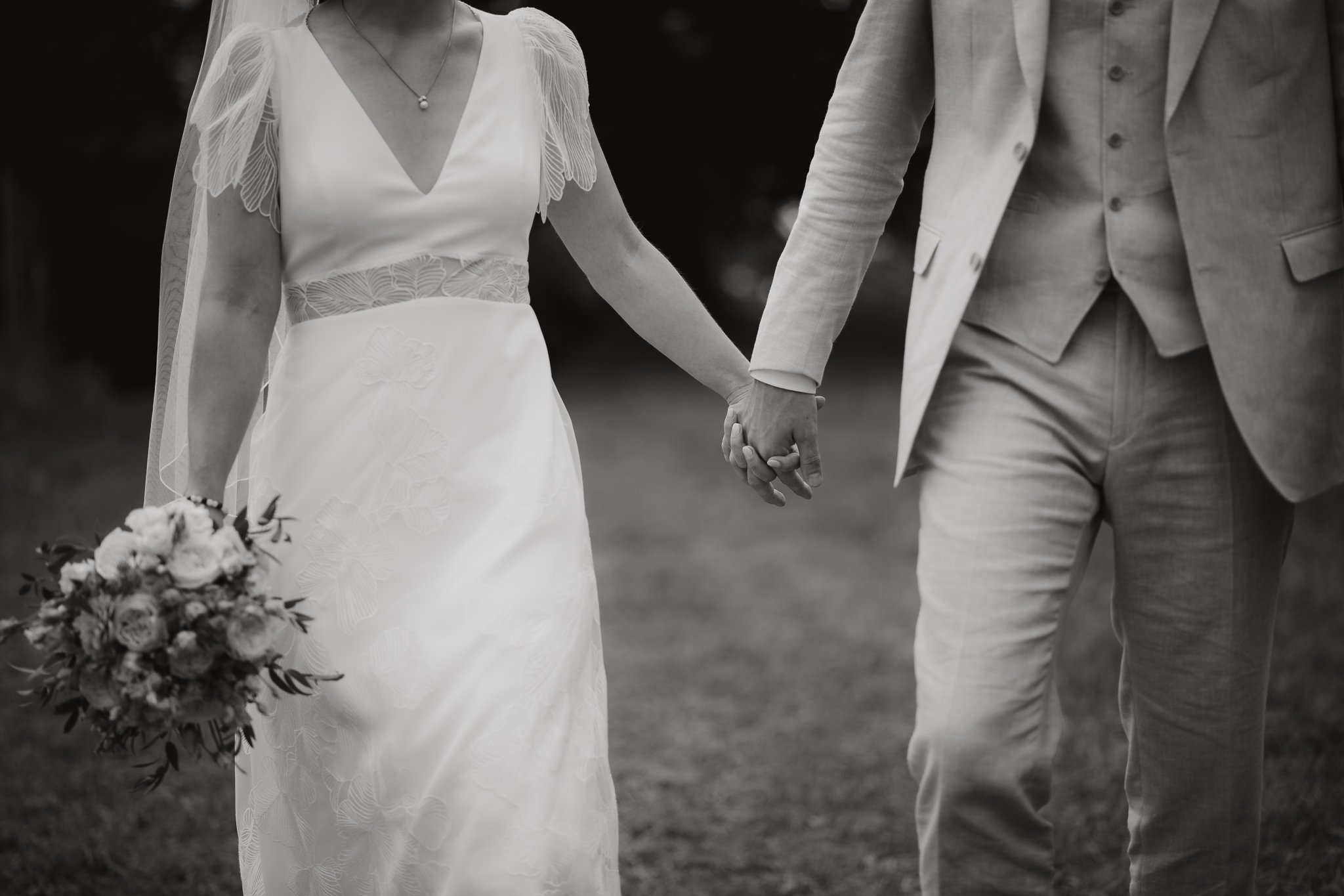 Ein Brautpaar hält sich an den Händen, die Braut trägt ein weißes Hochzeitskleid und eine Halskette, hält einen Blumenstrauß, der Bräutigam trägt einen hellen Anzug. Das Bild ist in Schwarzweiß gehalten.