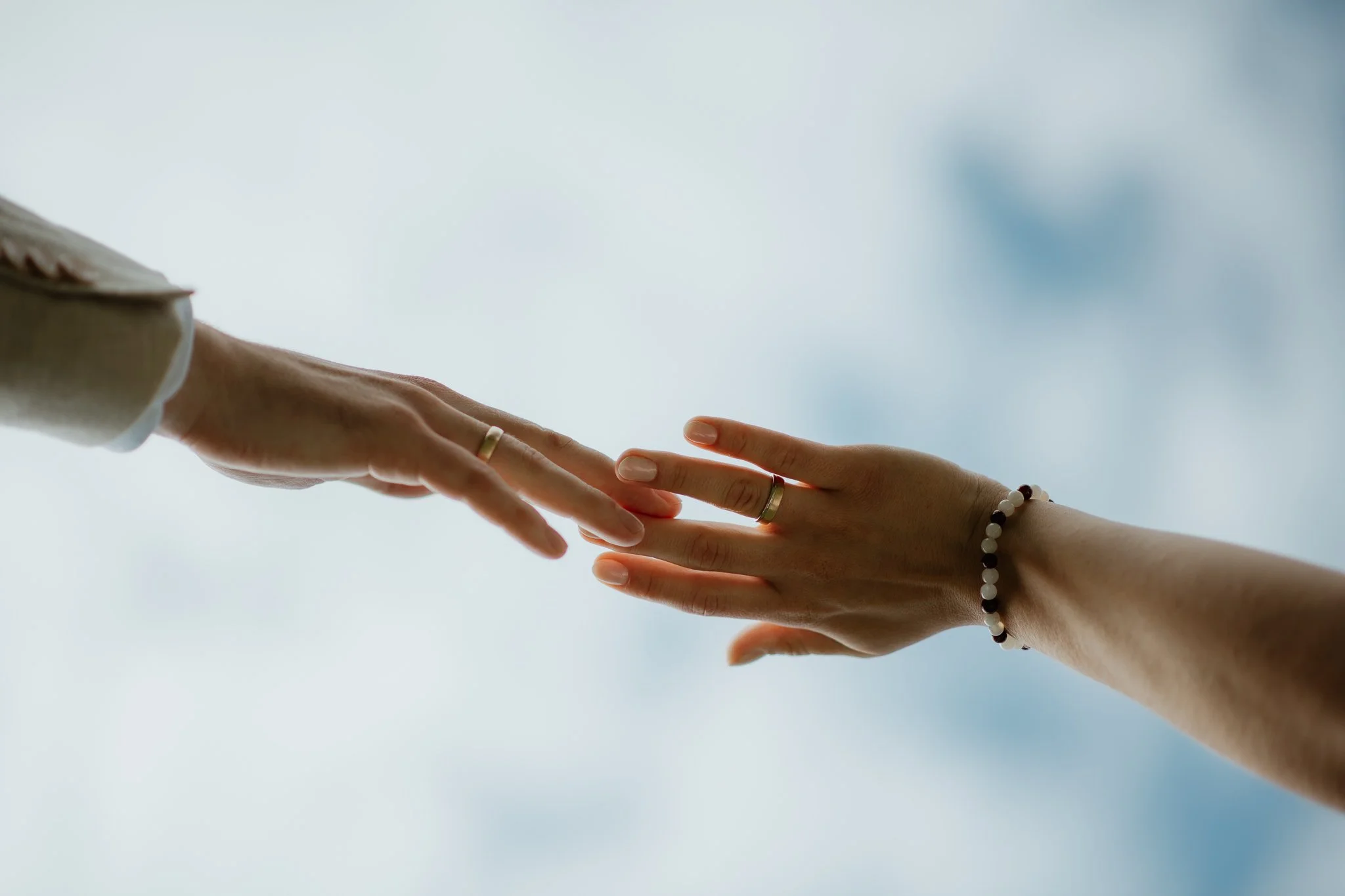 Nahaufnahme zweier Hände, die sich fast berühren, vor einem Himmel mit Wolken, beide tragen Ringe und eines hat eine Armbanduhr.