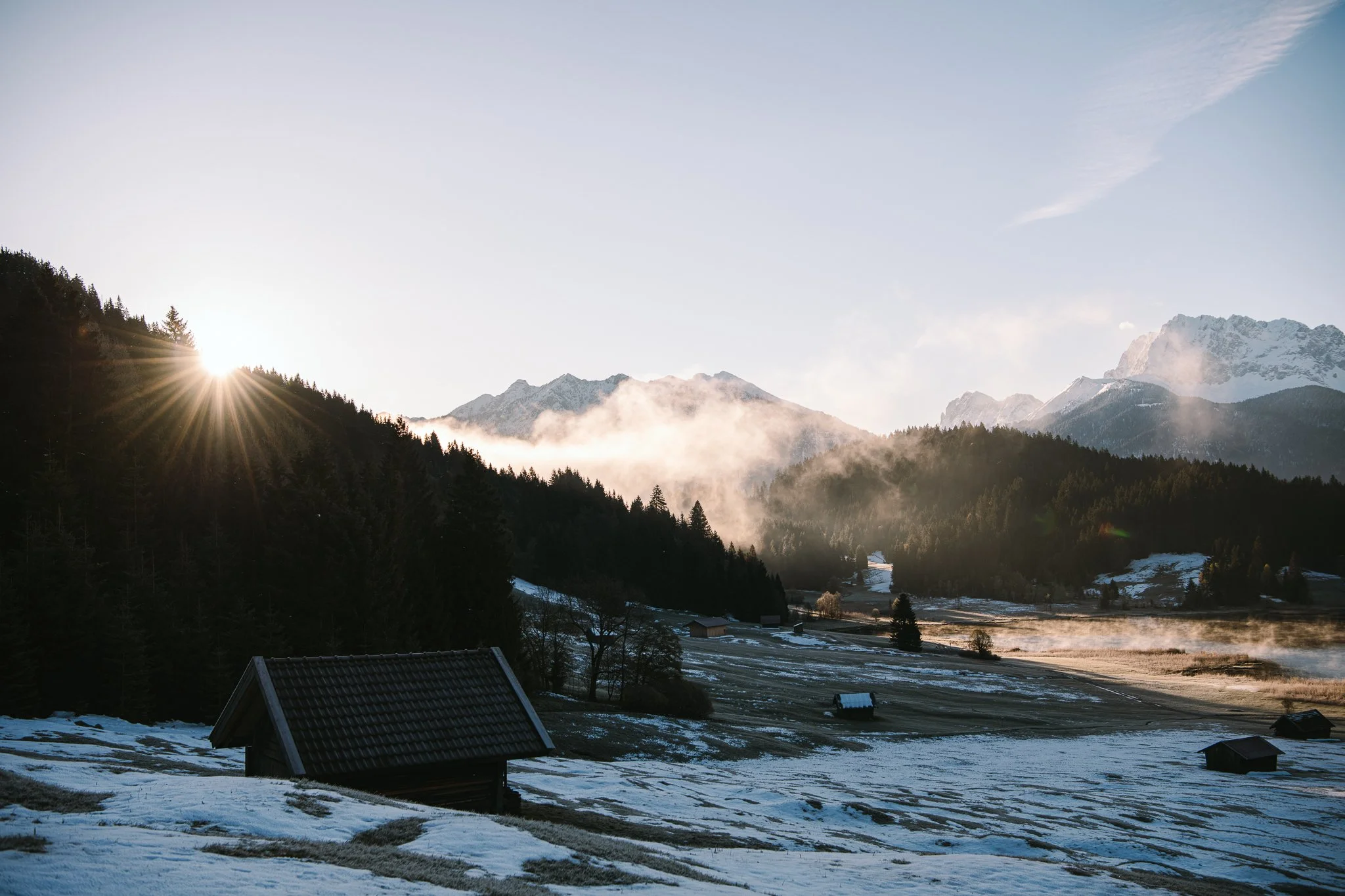 Berglandschaft mit Hütten und Sonne hinter Wald – Reisefotografie Garmisch Alpen
