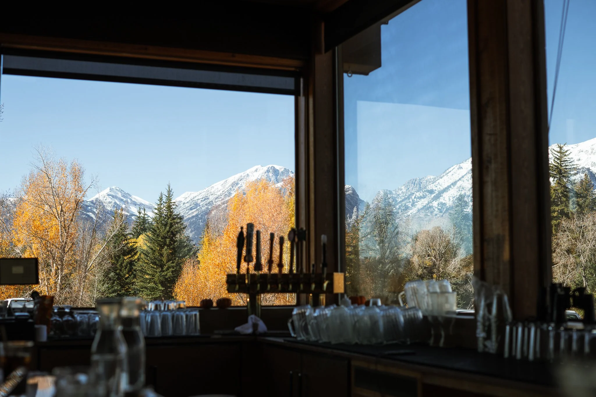 Blick aus einem Fenster mit Blick auf schneebedeckte Berge, Tannenbäume und Herbstbäume mit orangefarbenen Blätter, im Vordergrund eine Bar mit Gläsern und Flaschen.