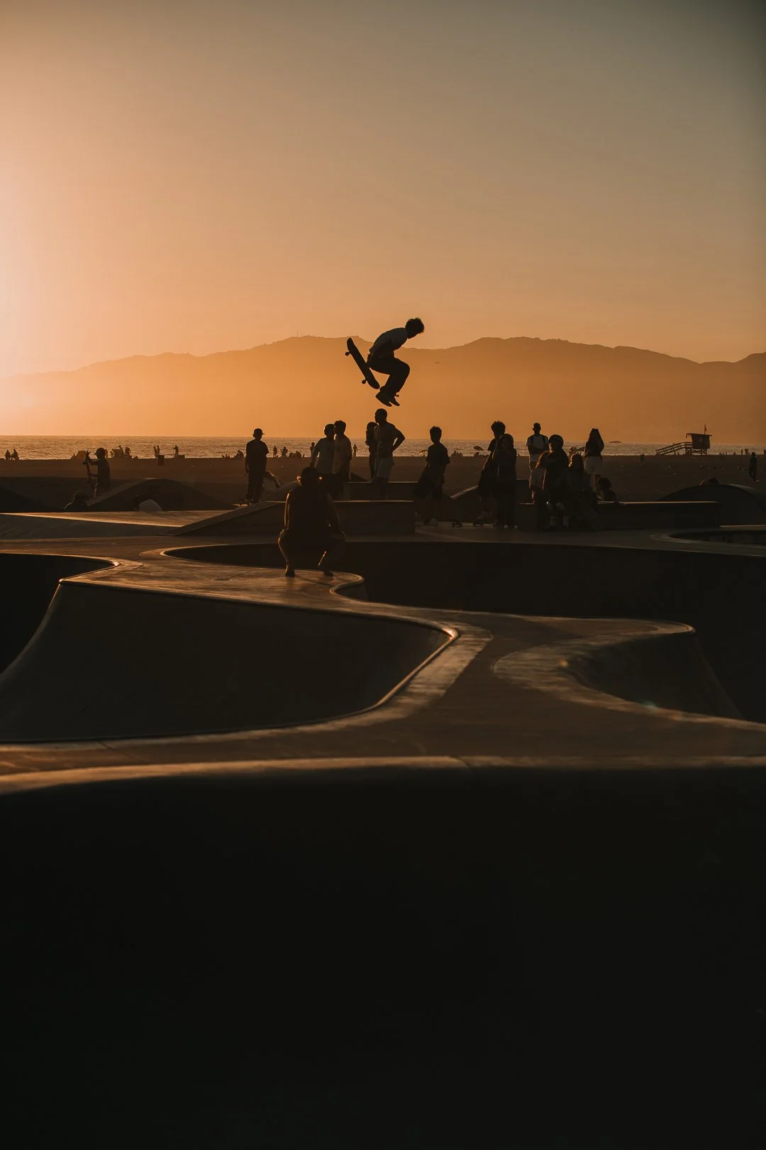 Ein Skateboarder springt in einem Skatepark bei Sonnenuntergang, während andere Leute im Hintergrund zuschauen.