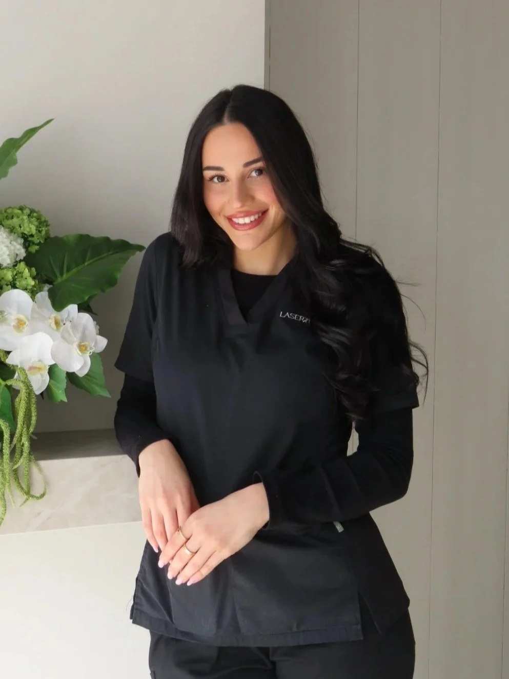 A woman with long dark hair smiling, wearing a black uniform with a Laser tattoo on her shirt, standing next to a large plant with white orchids and green leaves.